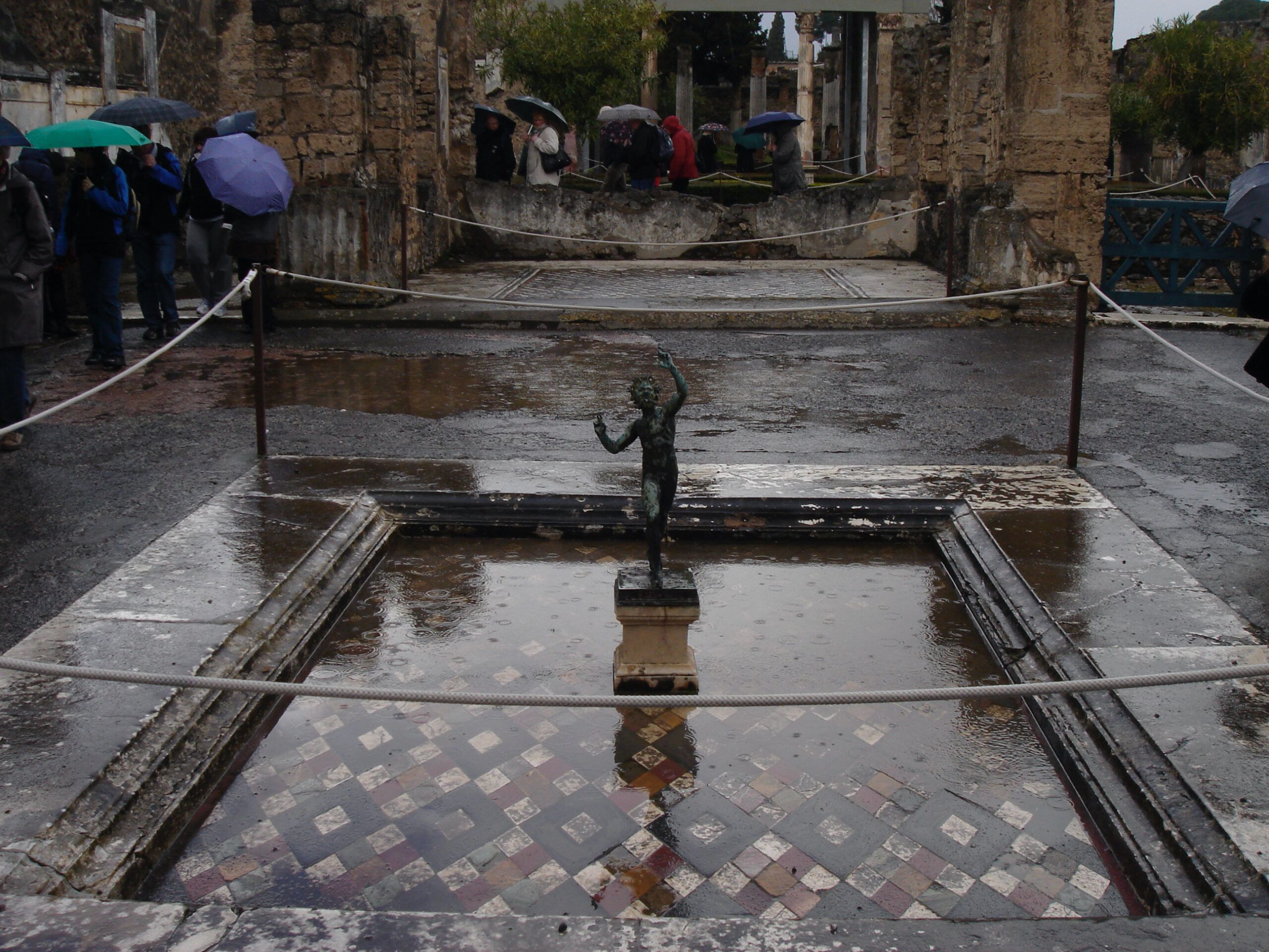 Rain-soaked atrium of the House of the Faun, Pompeii, with the famous bronze statue of a dancing faun standing in a tiled impluvium.