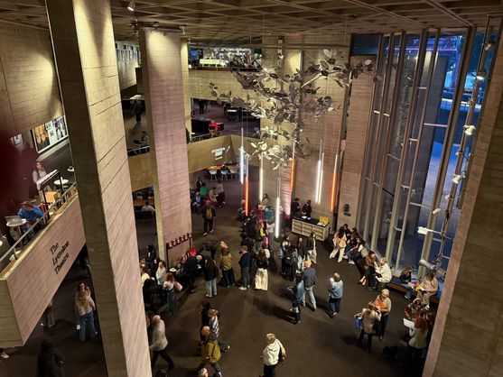 A wide view of the foyer of the National Theatre, with people milling about and sitting on benches and café tables. The space features tall pillars, large windows letting in evening light, and a hanging sculpture of metallic leaves.