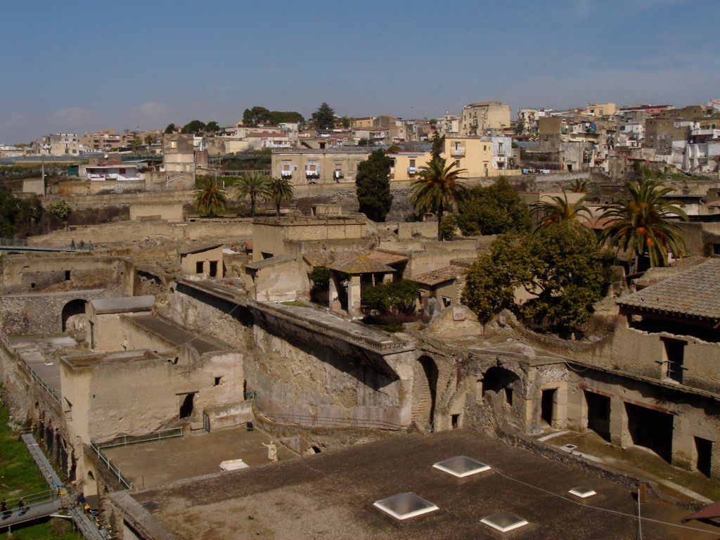View over the excavated ruins of Herculaneum with palm trees and the modern town rising in the background under a clear blue sky.