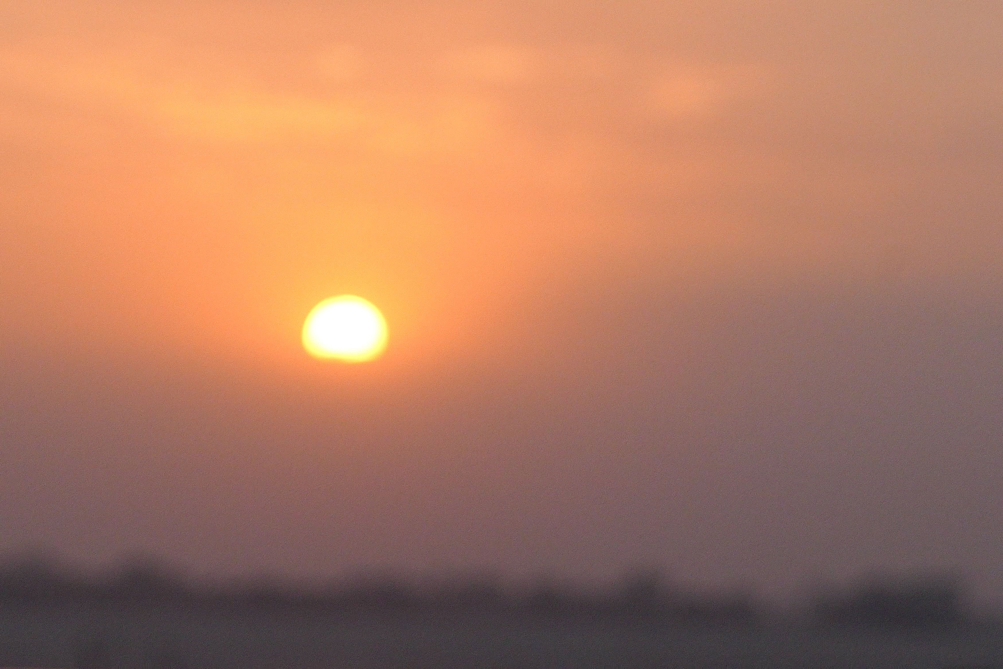 Hazy orange sun rises over the Ganges River, casting a warm glow over the distant horizon.