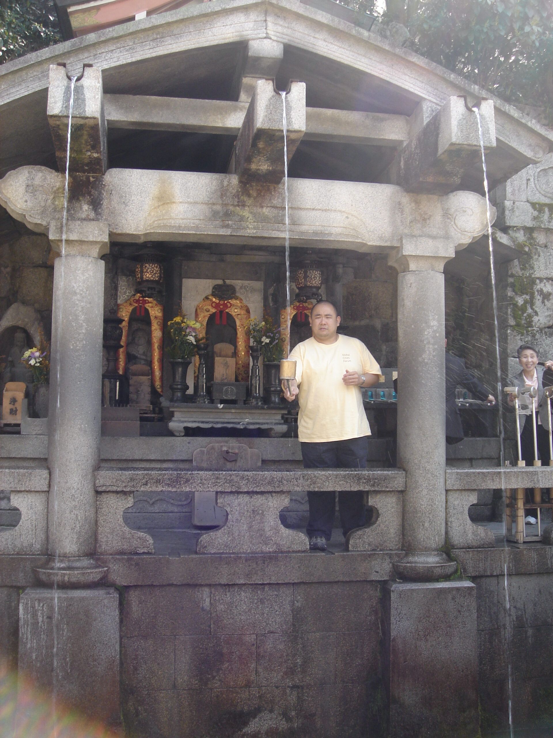 Calvin stands at a stone structure with three streams of water pouring down into cups, in front of small Buddhist statues with red bibs.
