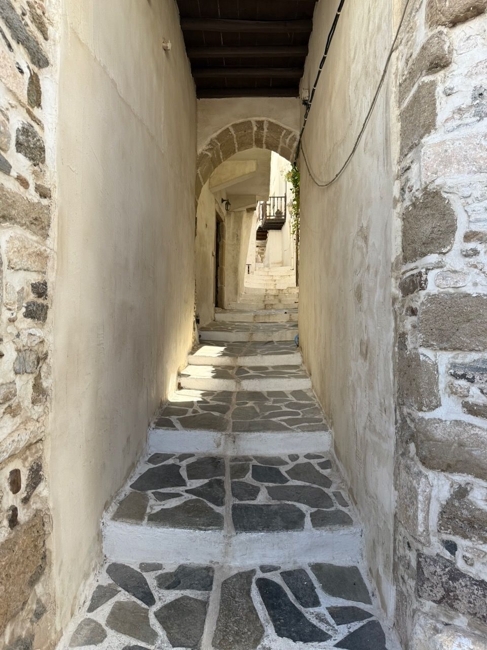 A narrow stone-paved staircase in the Kastro in Chora, Naxos, with whitewashed buildings on either side and an archway at the end.