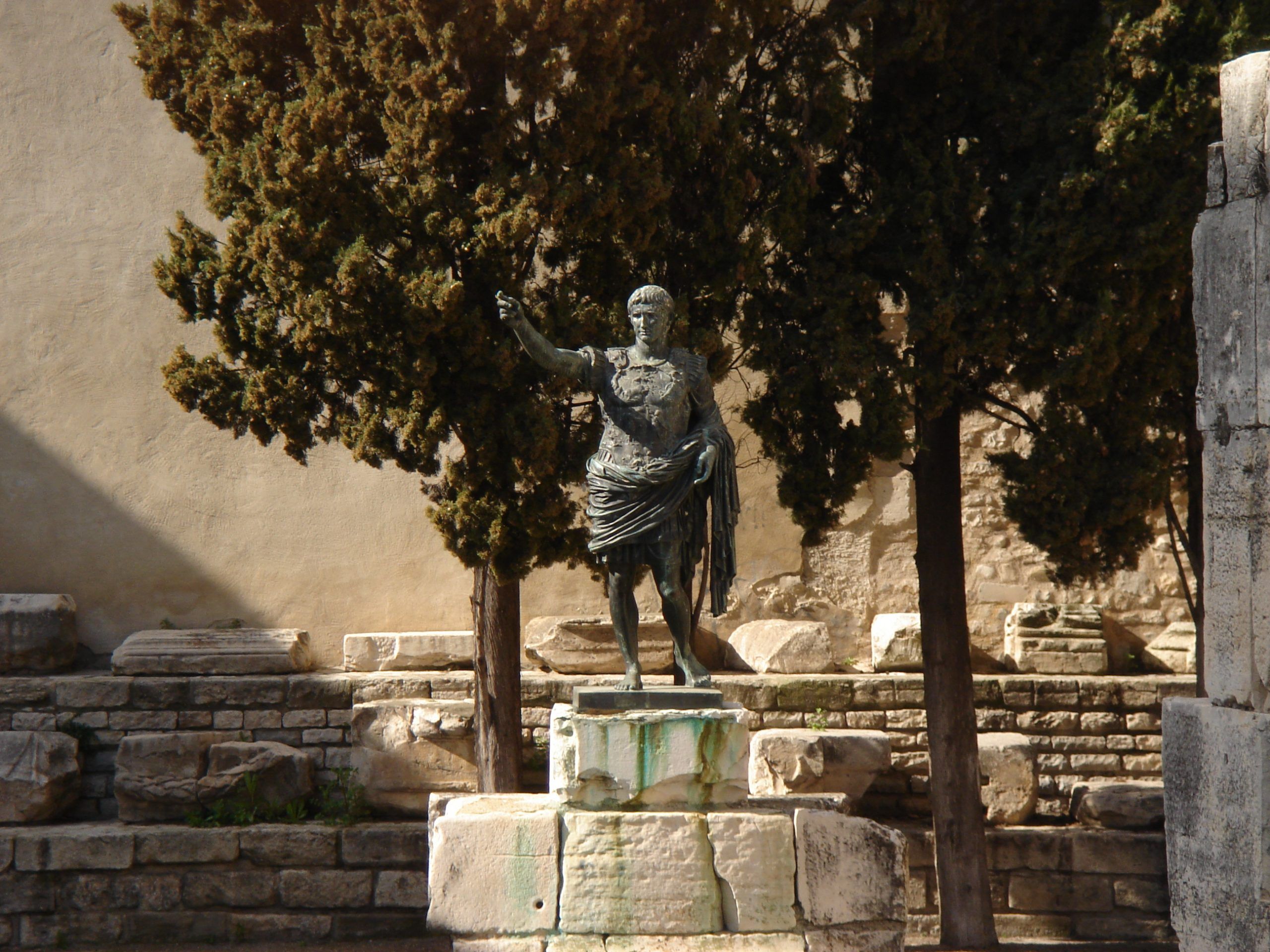 Bronze statue of a Roman figure in front of stone steps and cypress trees in the archaeological site at Nîmes.