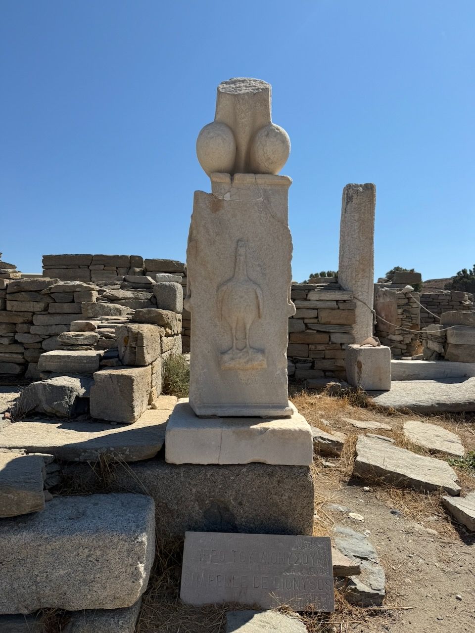 On a stone pedestal is an erect penis with balls. It has been broken off near the bottom of the shaft. On the front side of the pedestal is a rooster whose head has been replaced by an erect human penis.