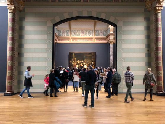 We're inside the Rijksmuseum in Amsterdam. Visitors are walking around in all directions. Behind them is a large arch in the wall with the name Rembrandt inscribed above it; behind that we can just make out The Night Watch.