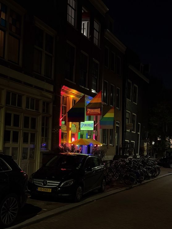 A street in Amsterdam at night. The front of the Spijkerbar is visible, lit up in rainbow lights and festooned with rainbow flags.