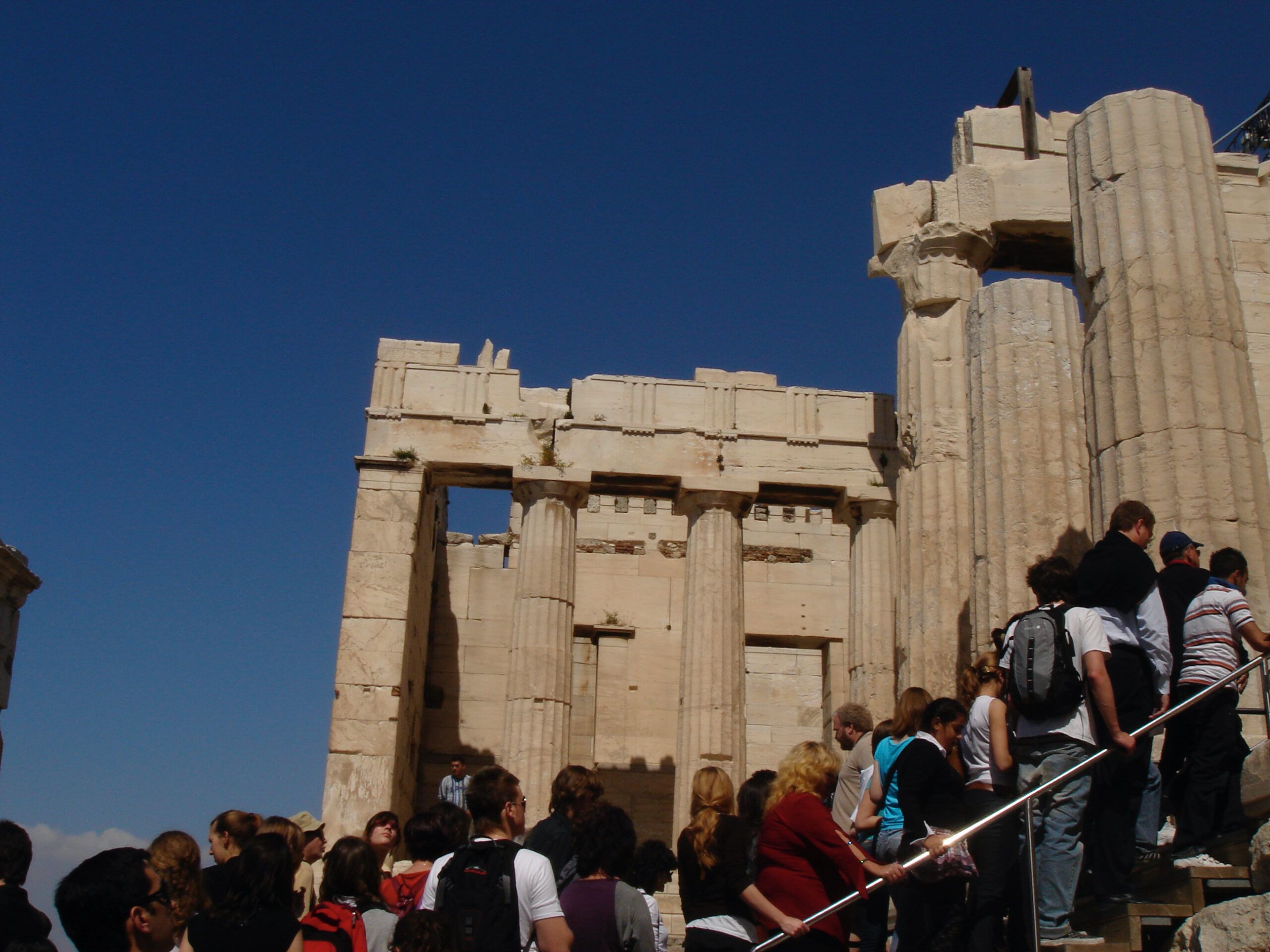 A crowd of tourists ascending the stone steps toward the Propylaia, the monumental gateway to the Acropolis in Athens, with large Doric columns and classical architecture visible under a clear blue sky.