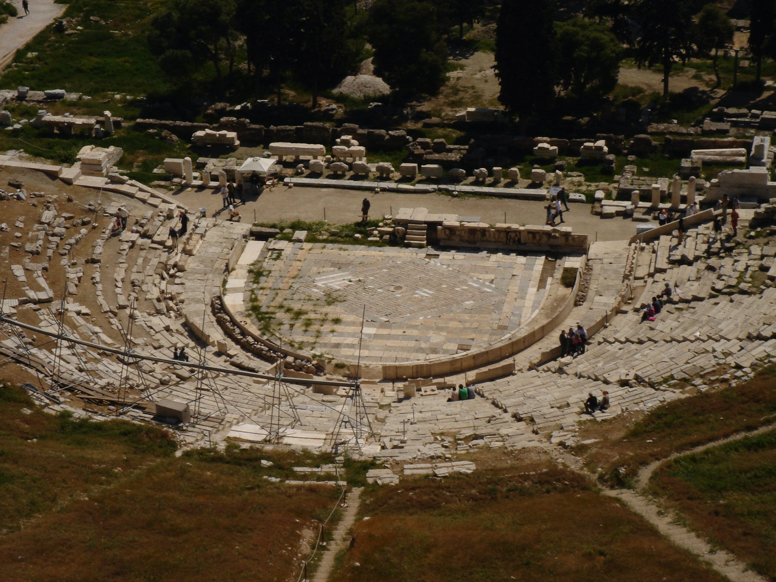 Overhead view of the ancient Theatre of Dionysus with partially restored seating and a central stage area, surrounded by scattered ruins.