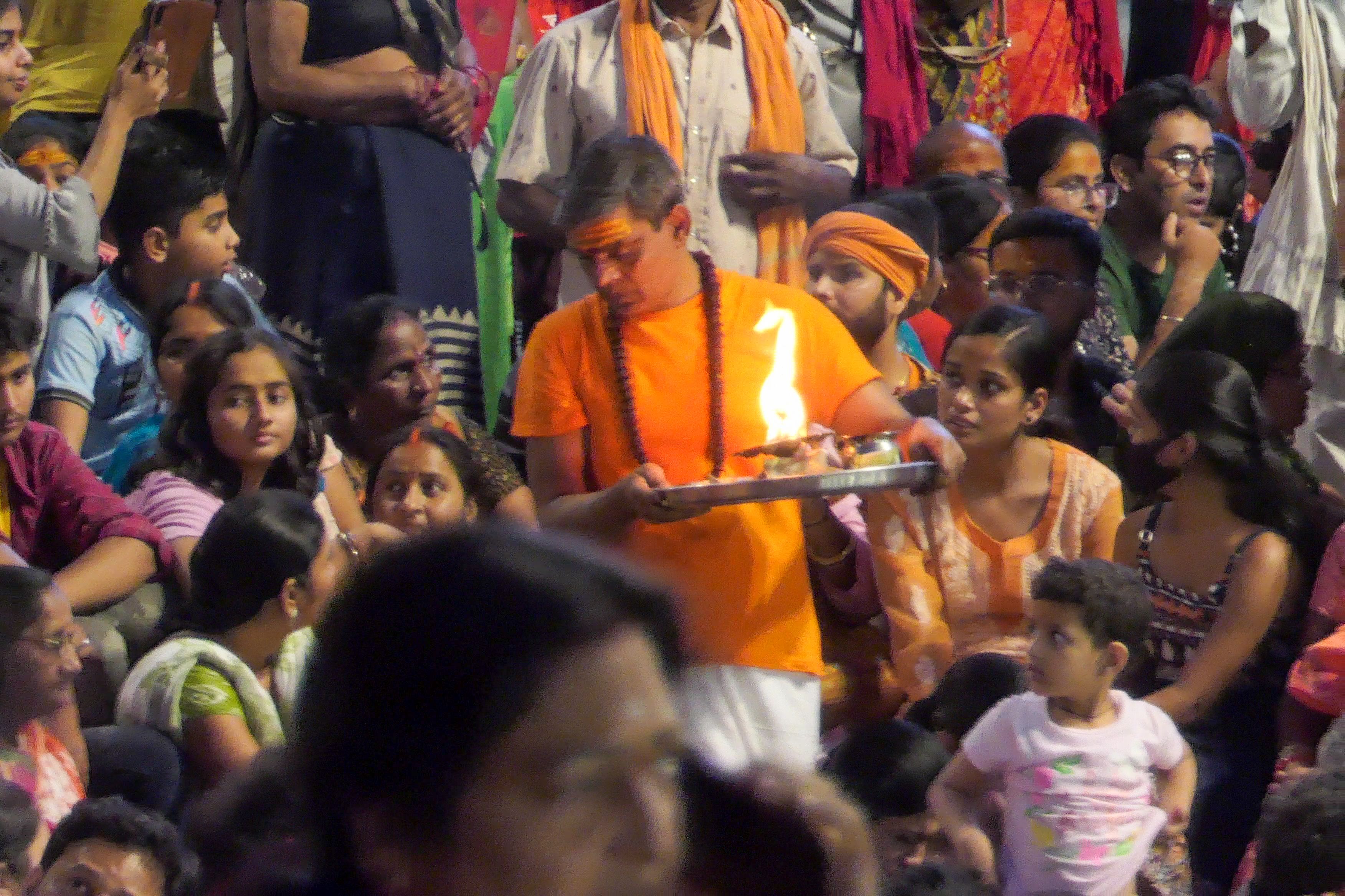 Devotee in an orange shirt holds a flaming aarti plate amidst a crowd at Dashashwamedh Ghat.