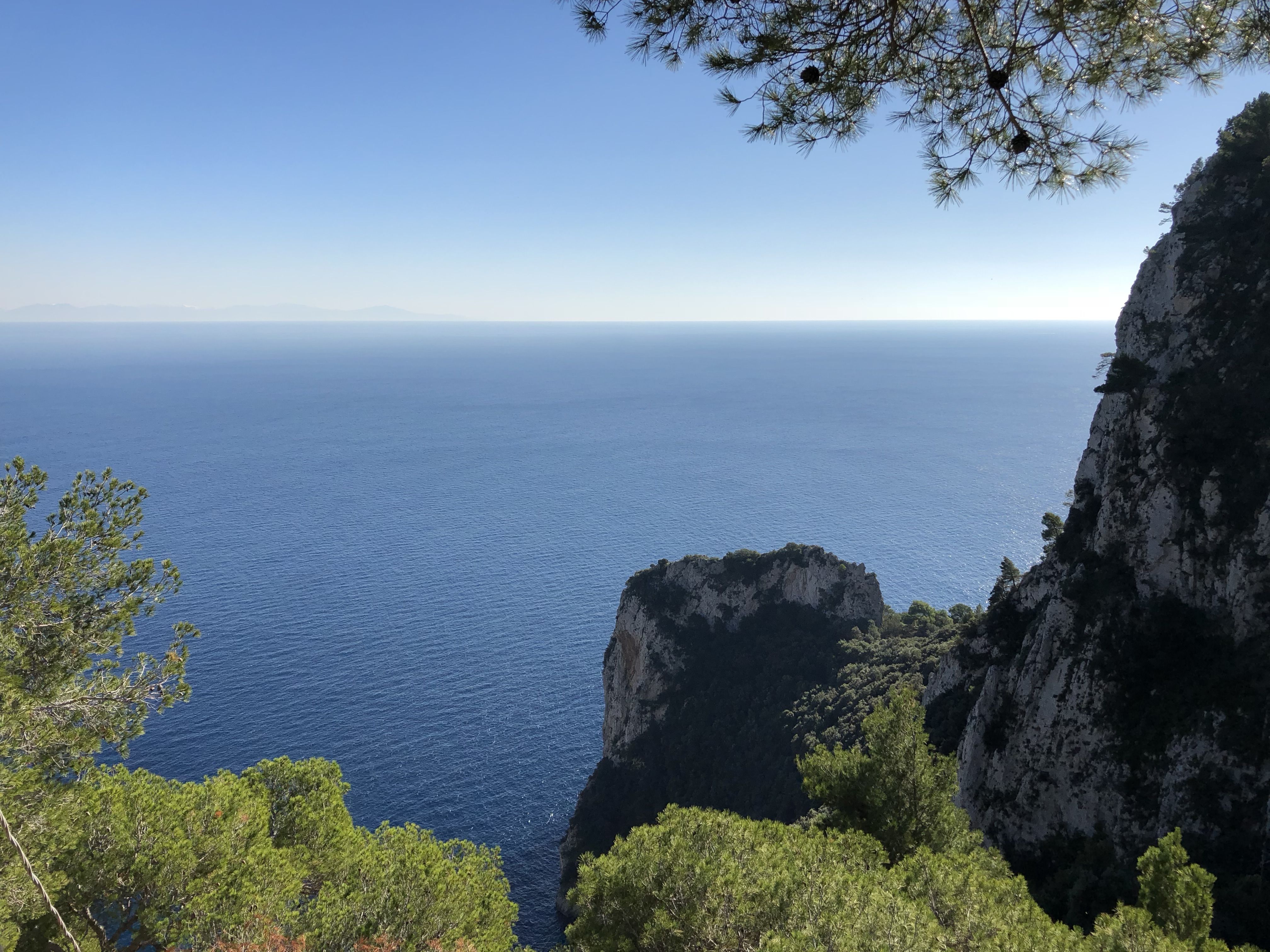 Vast expanse of blue water framed by trees and cliffs on Capri, looking out to the distant hazy mainland.