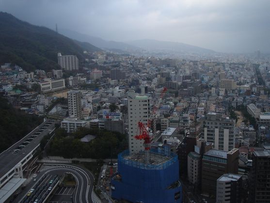 It's an overcast day and we're looking over the city of Kobe: mountains on the left, lots of multi-storey buildings clustered together, and just visible in the background on the right is the sea.