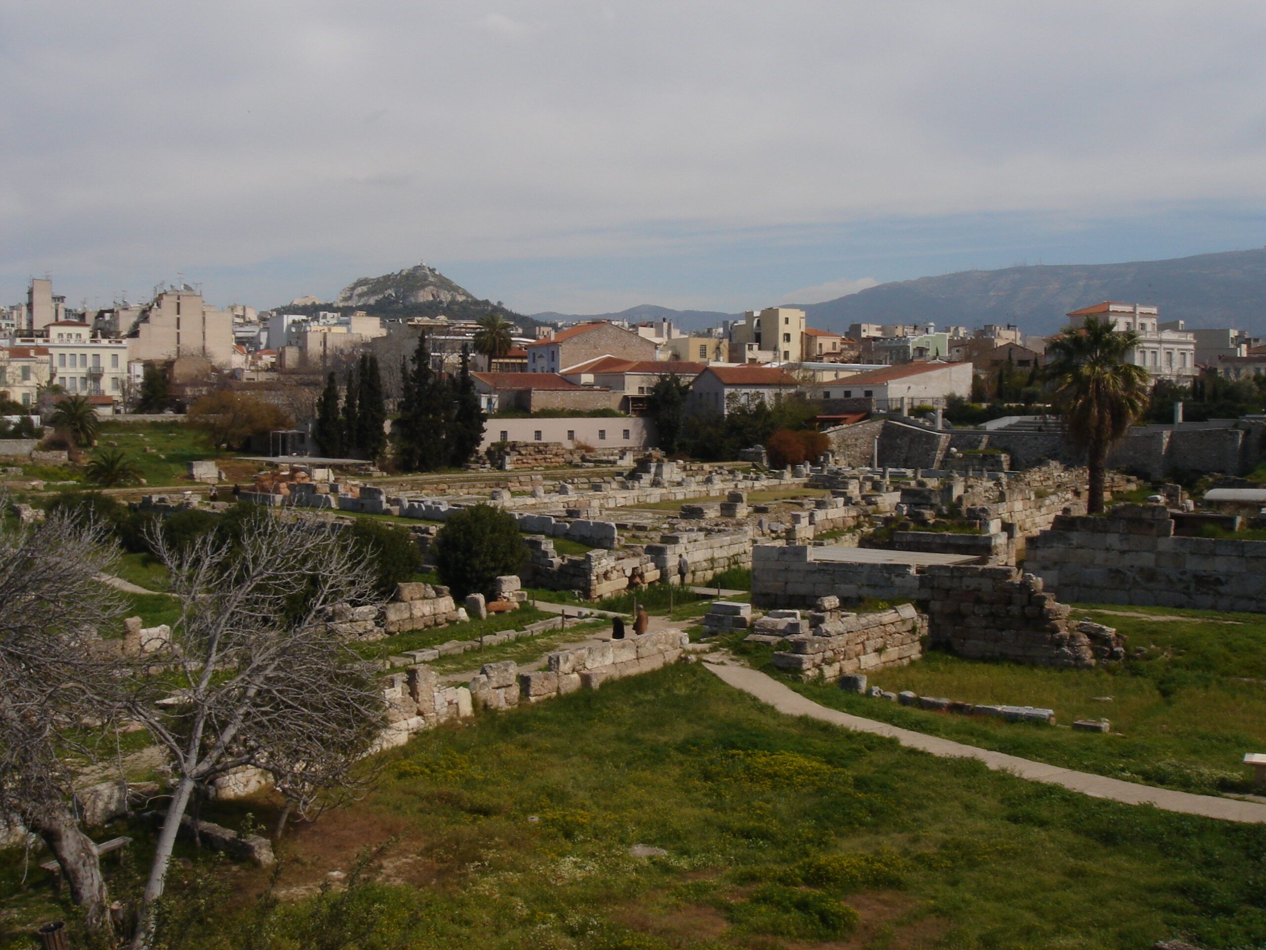 View across the ancient cemetery of the Kerameikos in Athens, with stone ruins scattered through green grass and modern buildings in the background.