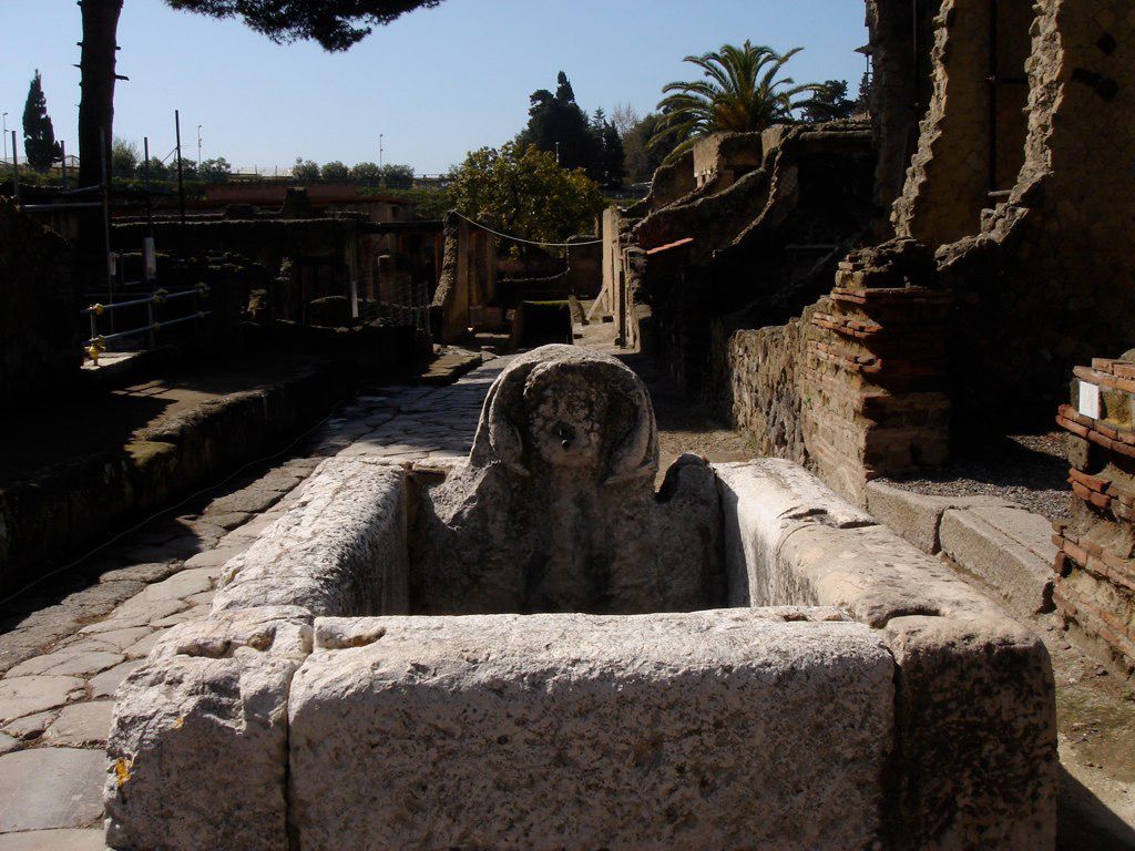 Stone fountain at a Herculaneum street corner, with a carved face spout and surrounding ruins, bathed in midday sunlight.