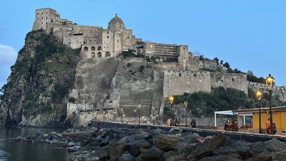 A hill juts out of the sea. There are walls built up the side facing us, and on top of those walls and the hill is a castle the same colour as the rock of the hill. A stone bridge leads from the bottom of the hill towards us.