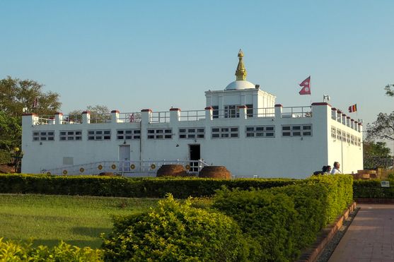 In a park a low building painted white, with a row of casement windows  below the roof. In the middle of the roof is a little tower with a dome on  top with a golden pillar on top. There's a walkway in front of it with an open door.
