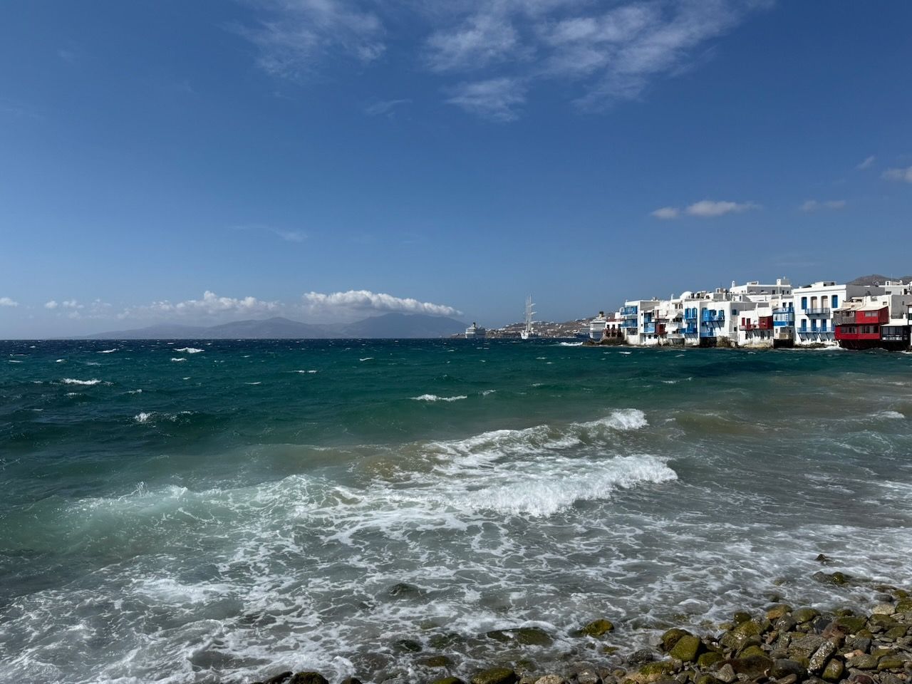 The seaside, with breaking waves and a row of colourful shops and houses and restaurants in the distance. Beyond them a cruise ship and a boat with three masts.