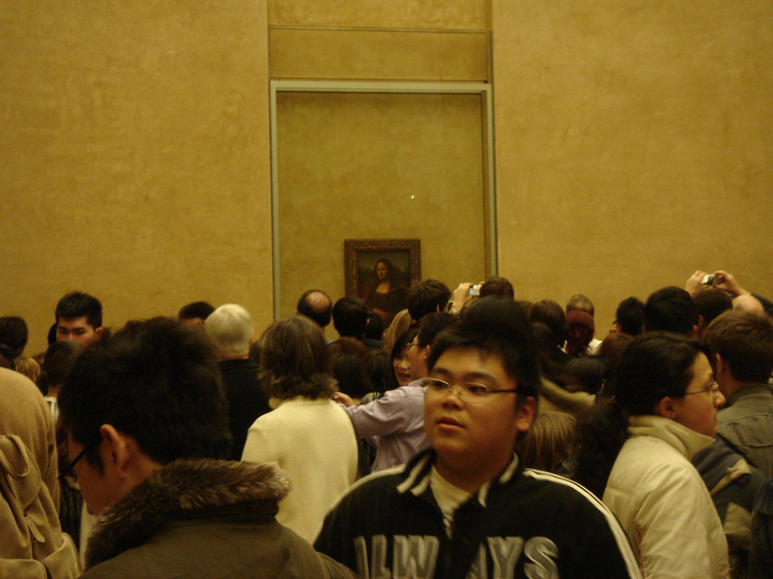 Large crowd gathered inside the Louvre Museum viewing the Mona Lisa behind protective glass.