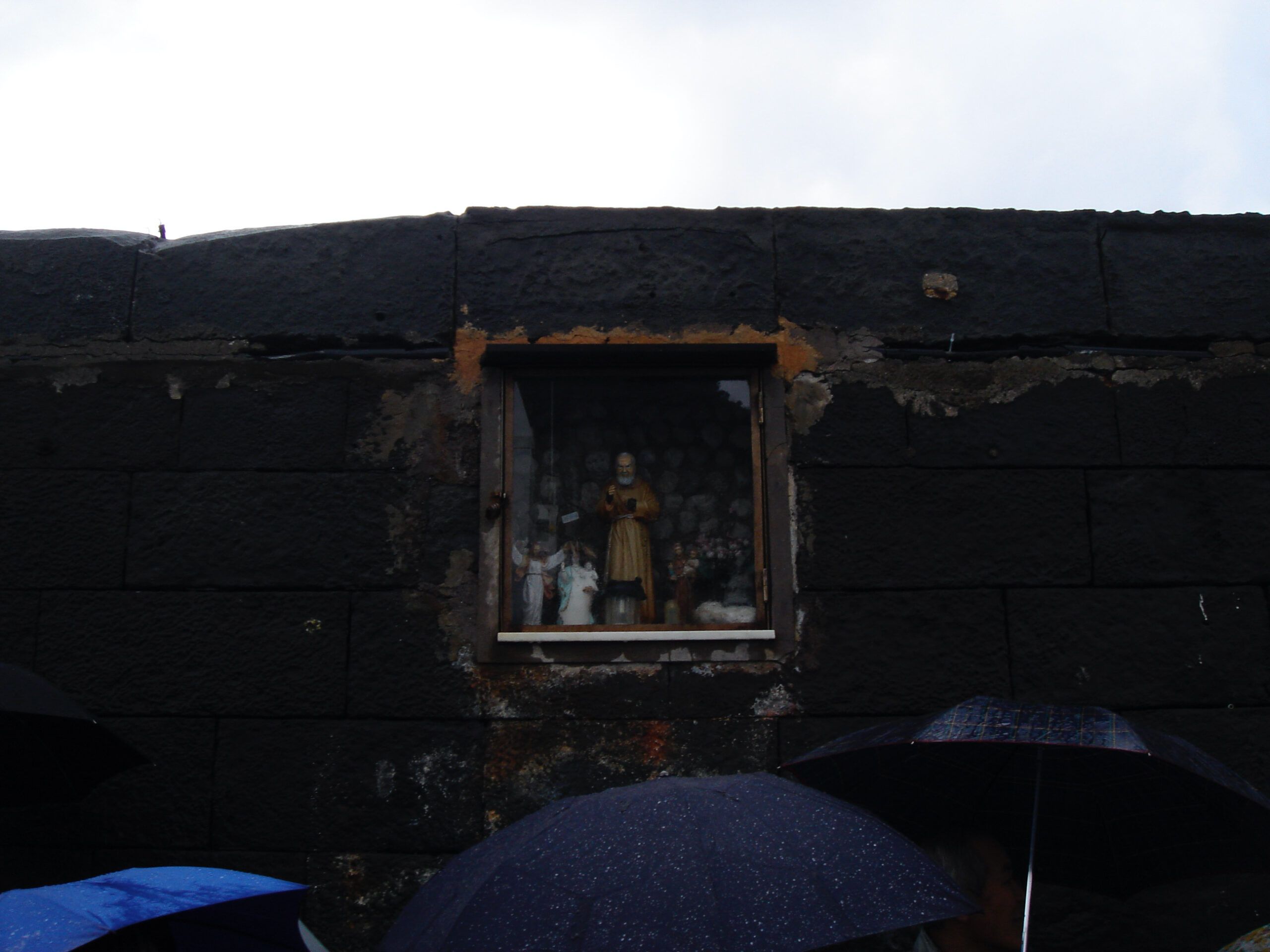 A small shrine built into a dark stone wall, featuring a religious statue behind glass, with people holding umbrellas below.