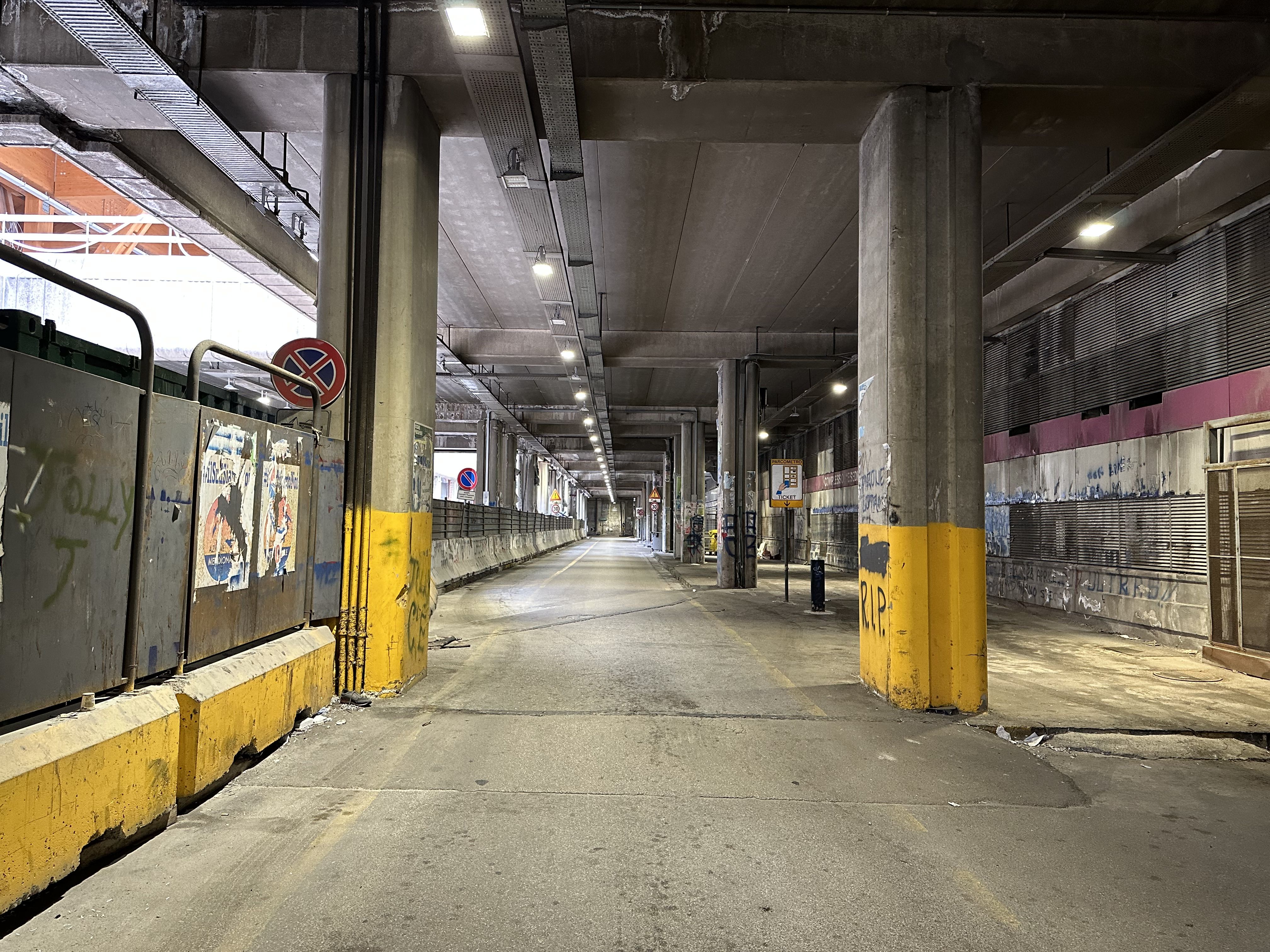 Dimly lit, graffiti-covered parking structure in Naples with concrete pillars and litter-strewn floor.