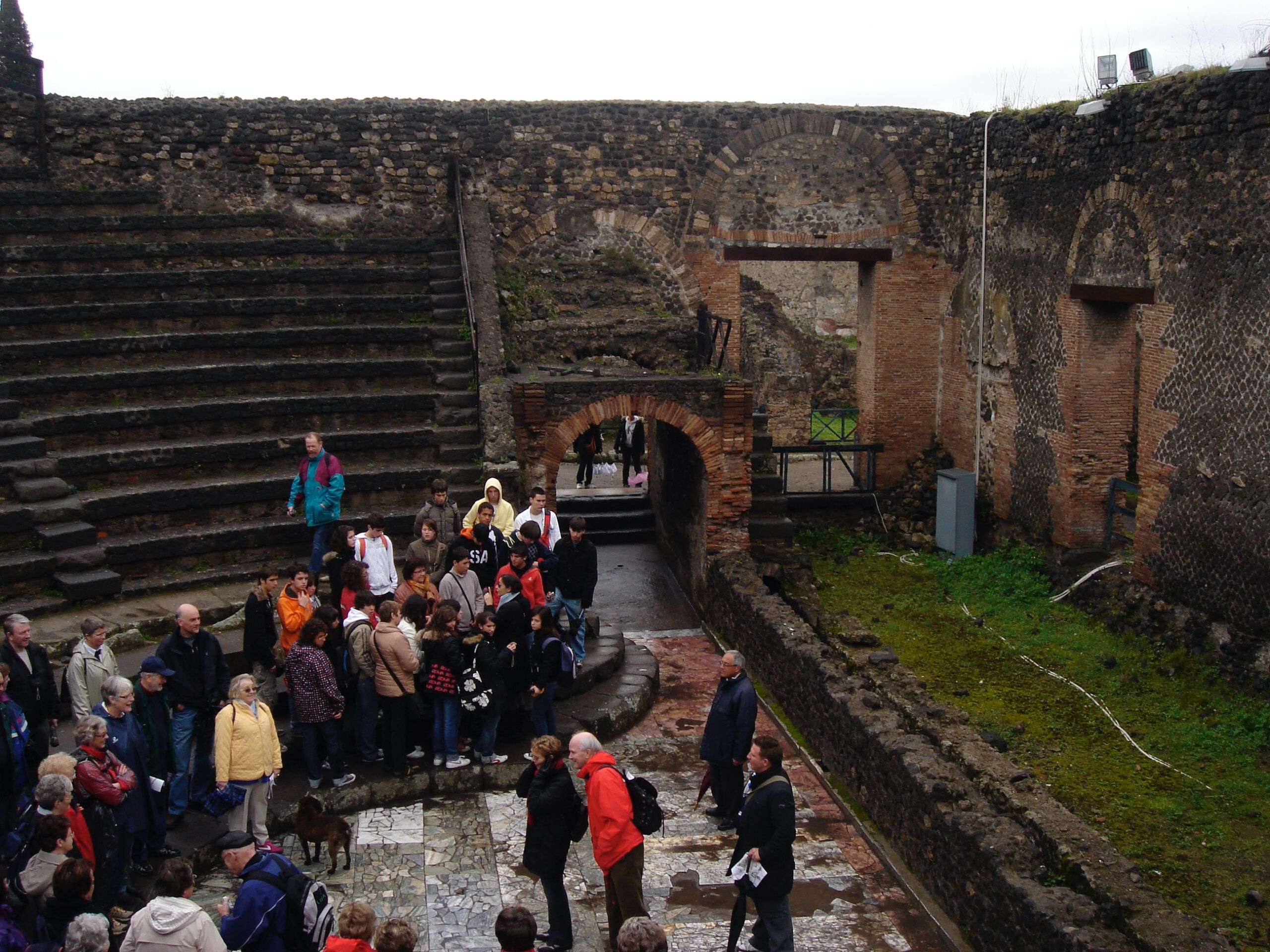 Tour group gathered in the lower seating area of the small theatre at Pompeii, surrounded by arched brick walls and mosaic flooring.