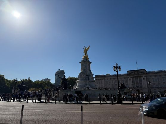 The Victoria Memorial with its gilded statue shining in the sunlight in front of Buckingham Palace, London, on a clear blue day with crowds gathered around the monument.