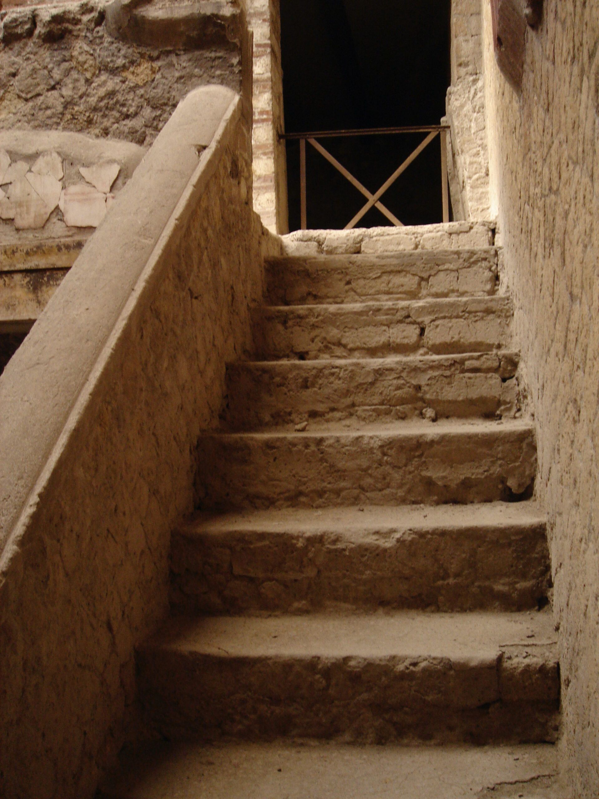 Narrow staircase in Herculaneum with worn stone steps leading up to a shadowed doorway and iron railing at the top.
