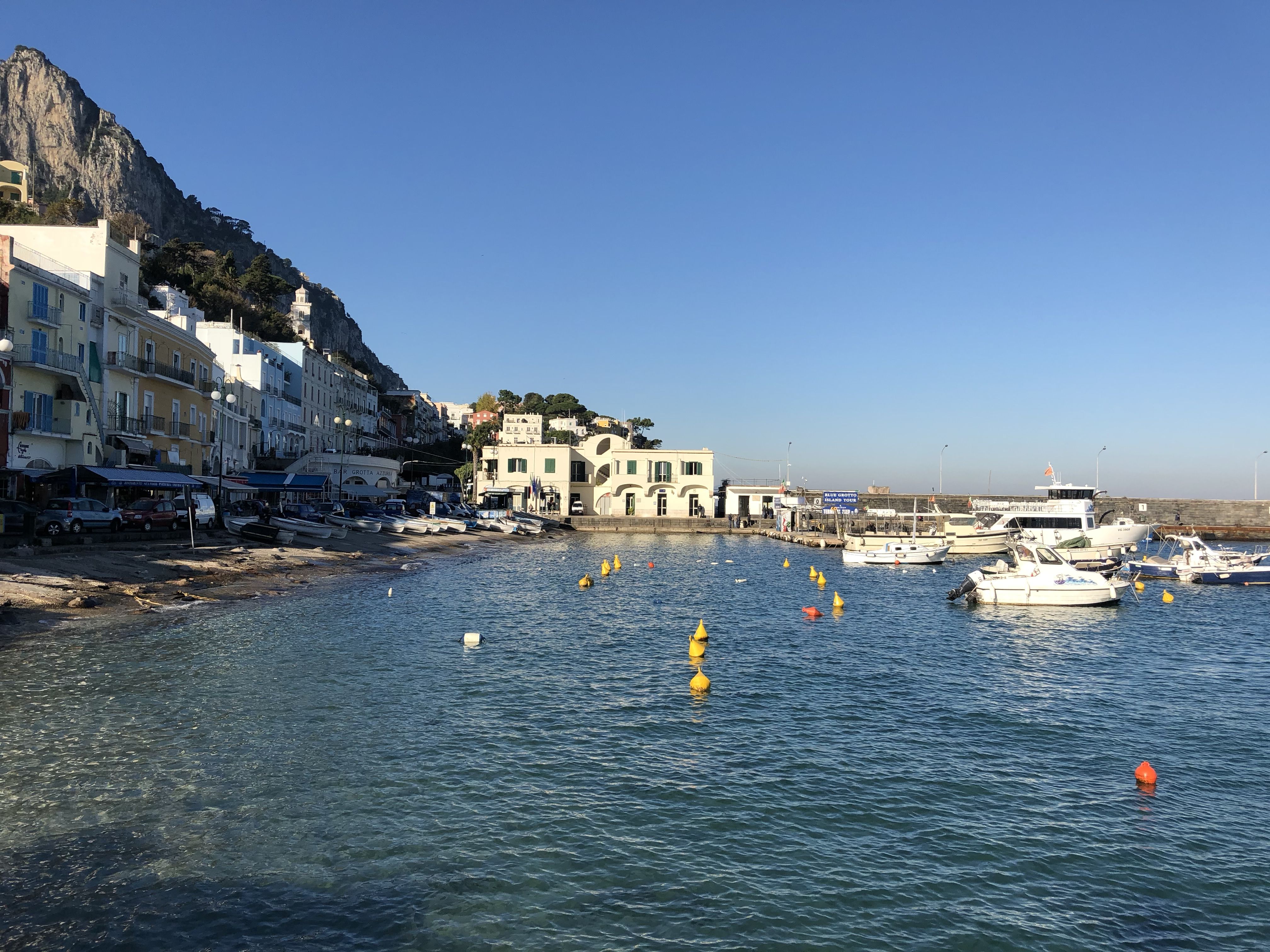 Boats float near the marina of Capri, with colourful buildings lining the waterfront and steep cliffs rising behind.