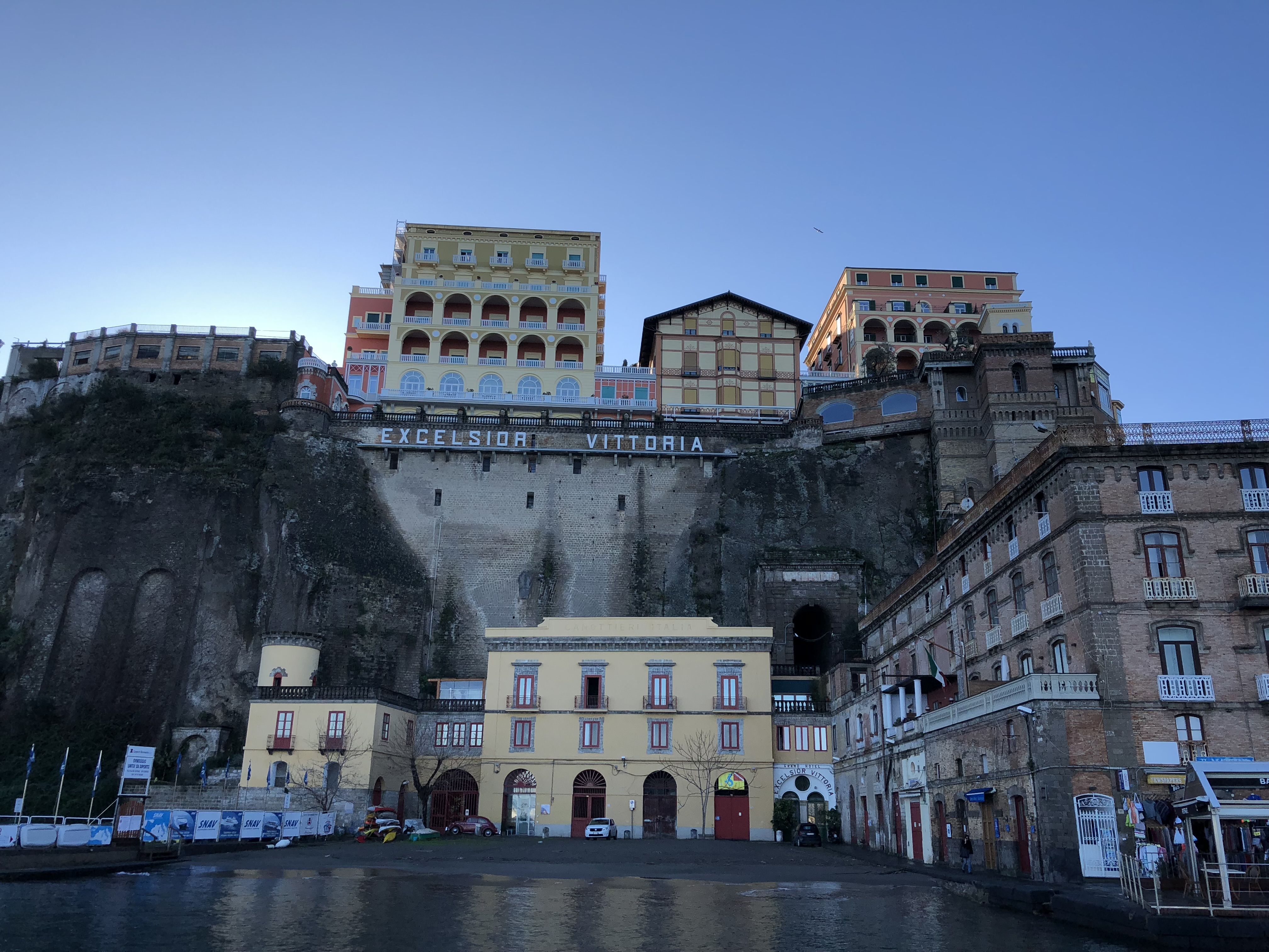 There's a cliff directly ahead of us, no, it's a brick wall with some old brick arches and water dripping down the surface. On the top are the words Excelsior Vittoria, and above them are two brightly-coloured hotels. Below, is the Excelsior Vittoria itself, a hotel painted pale yellow.