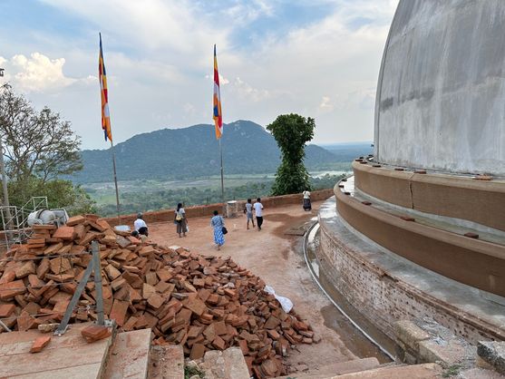 A group of pilgrims walks around the base of a large white stupa under restoration, with piles of bricks nearby and Buddhist flags flying above a panoramic mountain view at Mihintale.