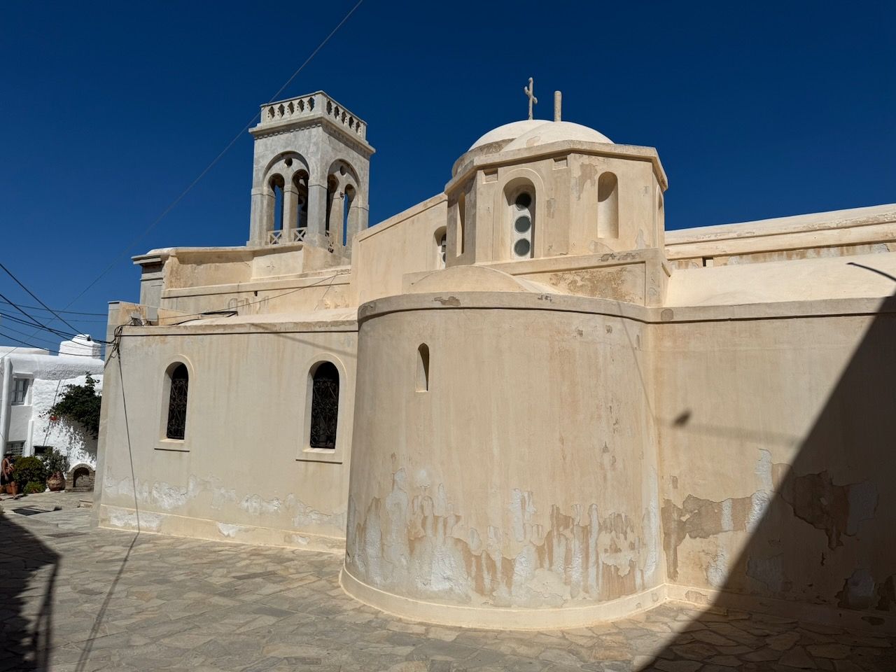 The Catholic Catheral in Naxos, a beige-plastered building with a domed apse and tall arched bell tower, set in the narrow stone-paved lanes of the Kastro in Chora, Naxos under a deep blue sky.
