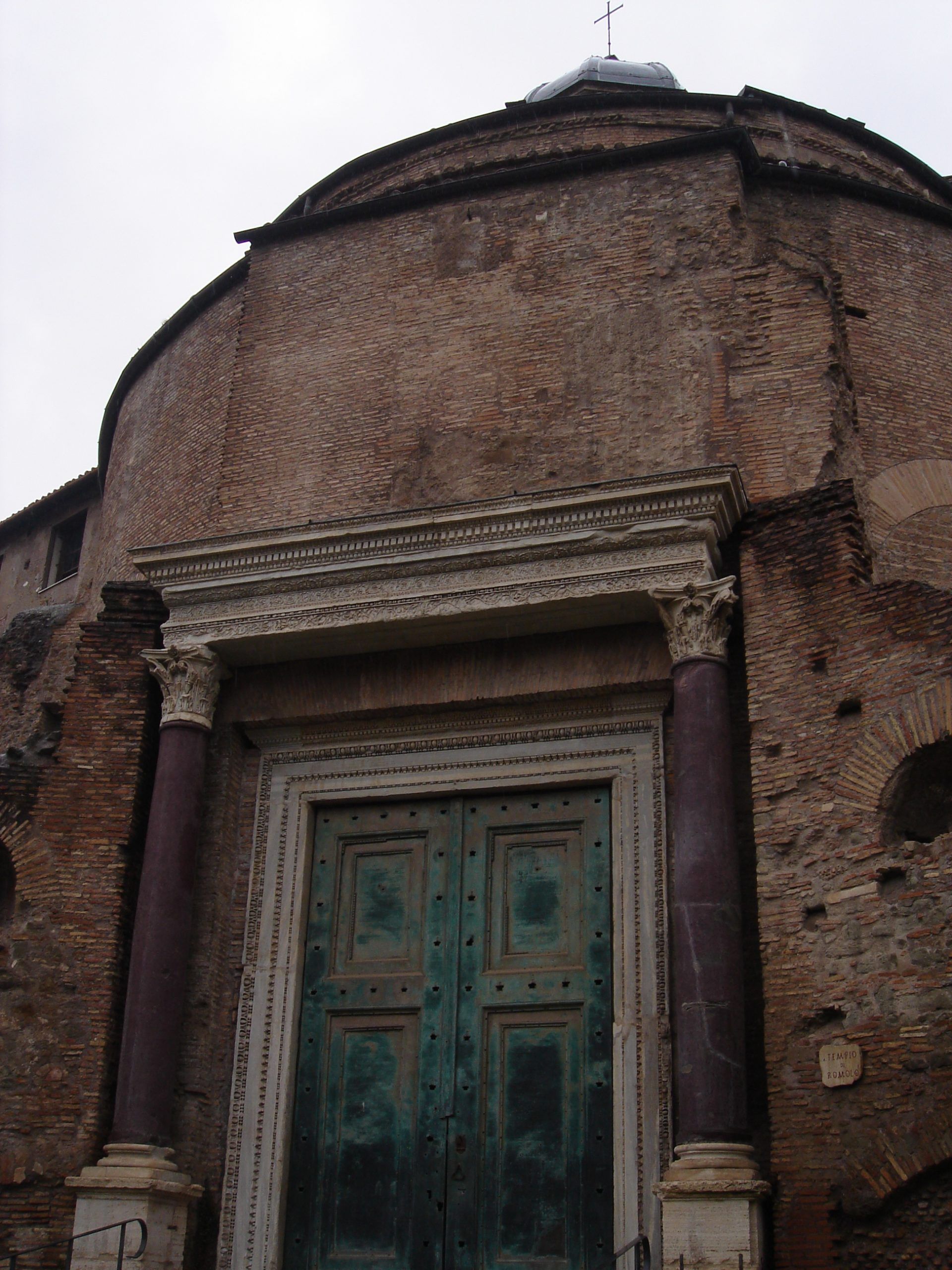 Close-up of the ancient bronze doors of the Temple of Romulus in the Roman Forum, framed by purple porphyry columns and a worn brick rotunda.
