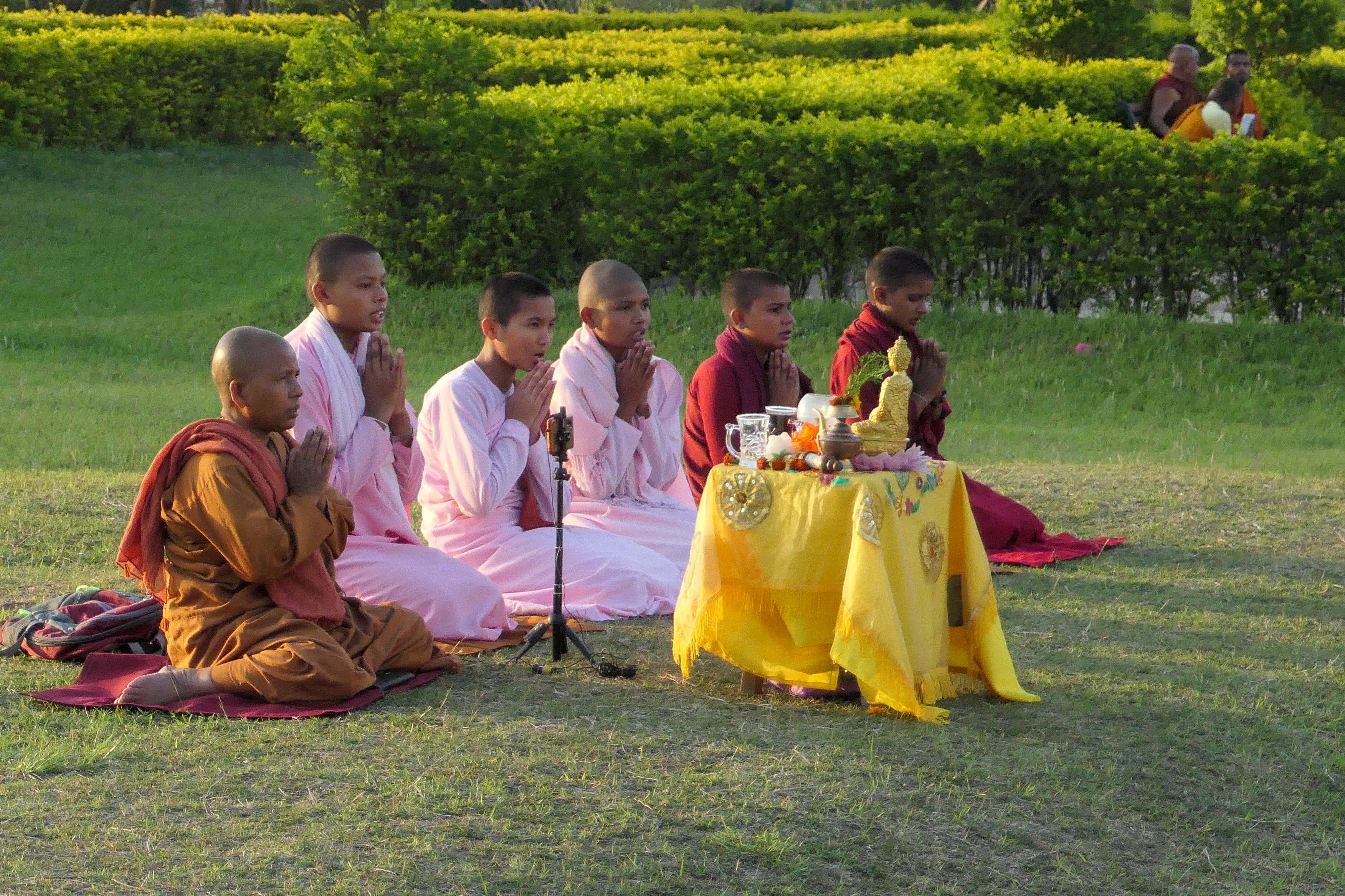 Group of young monks and nuns in colorful robes kneeling in prayer around a small altar with a golden Buddha statue at Lumbinī.