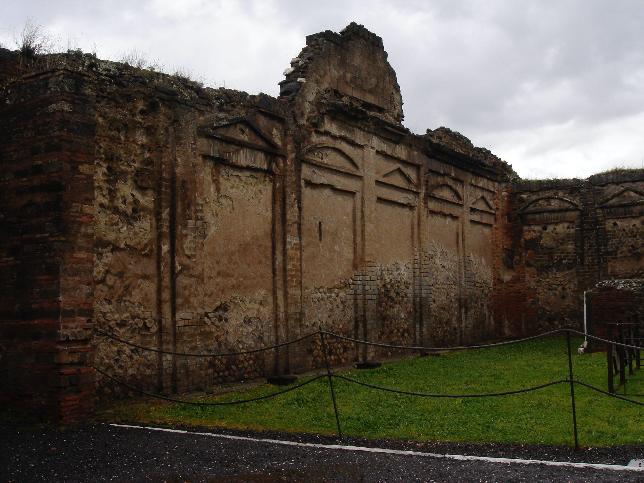 A large stone wall with faded decorative reliefs stands on the grassy edge of Pompeii's Forum near the Temple of Jupiter.