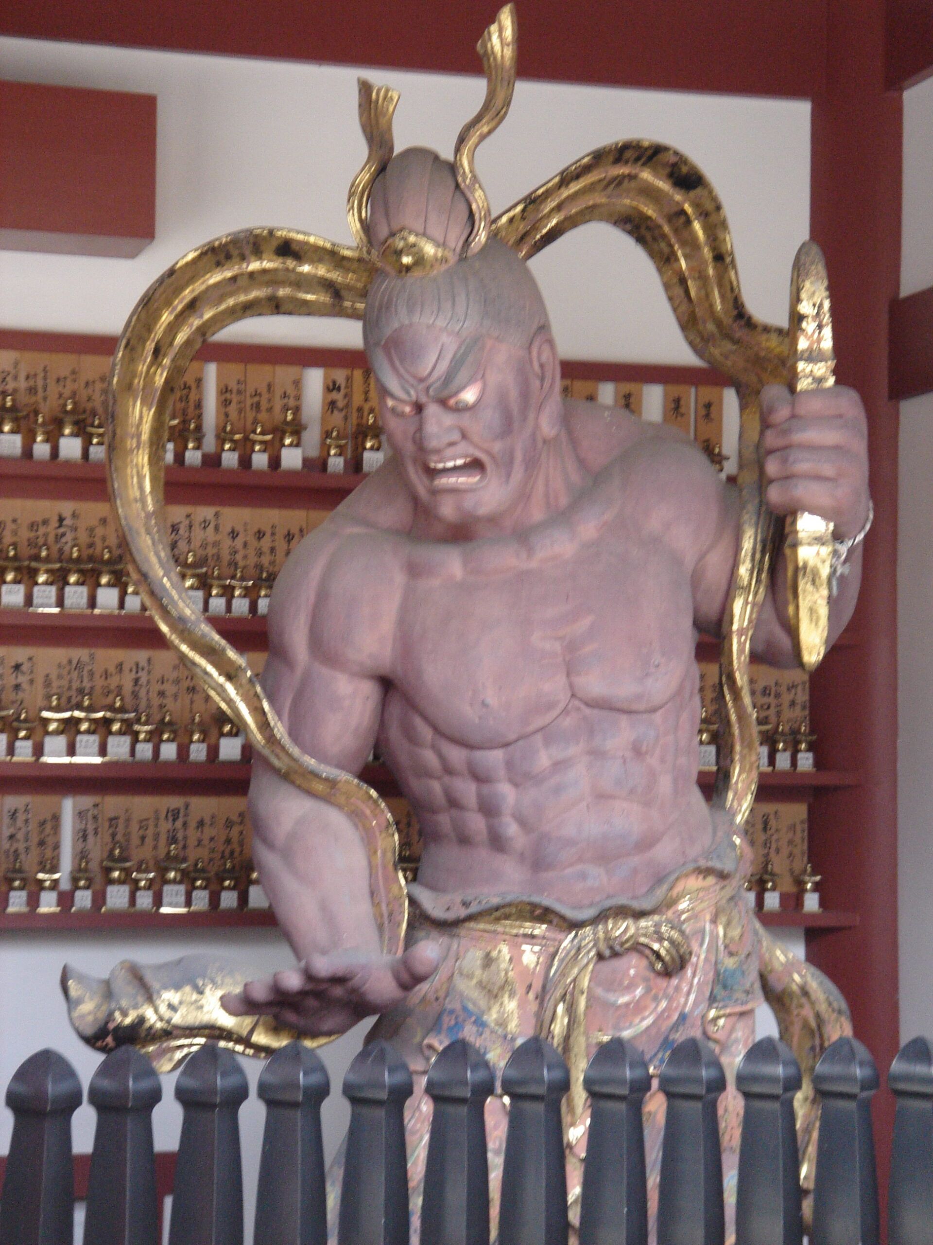 A fierce guardian statue with muscular form and gold accents inside a temple.