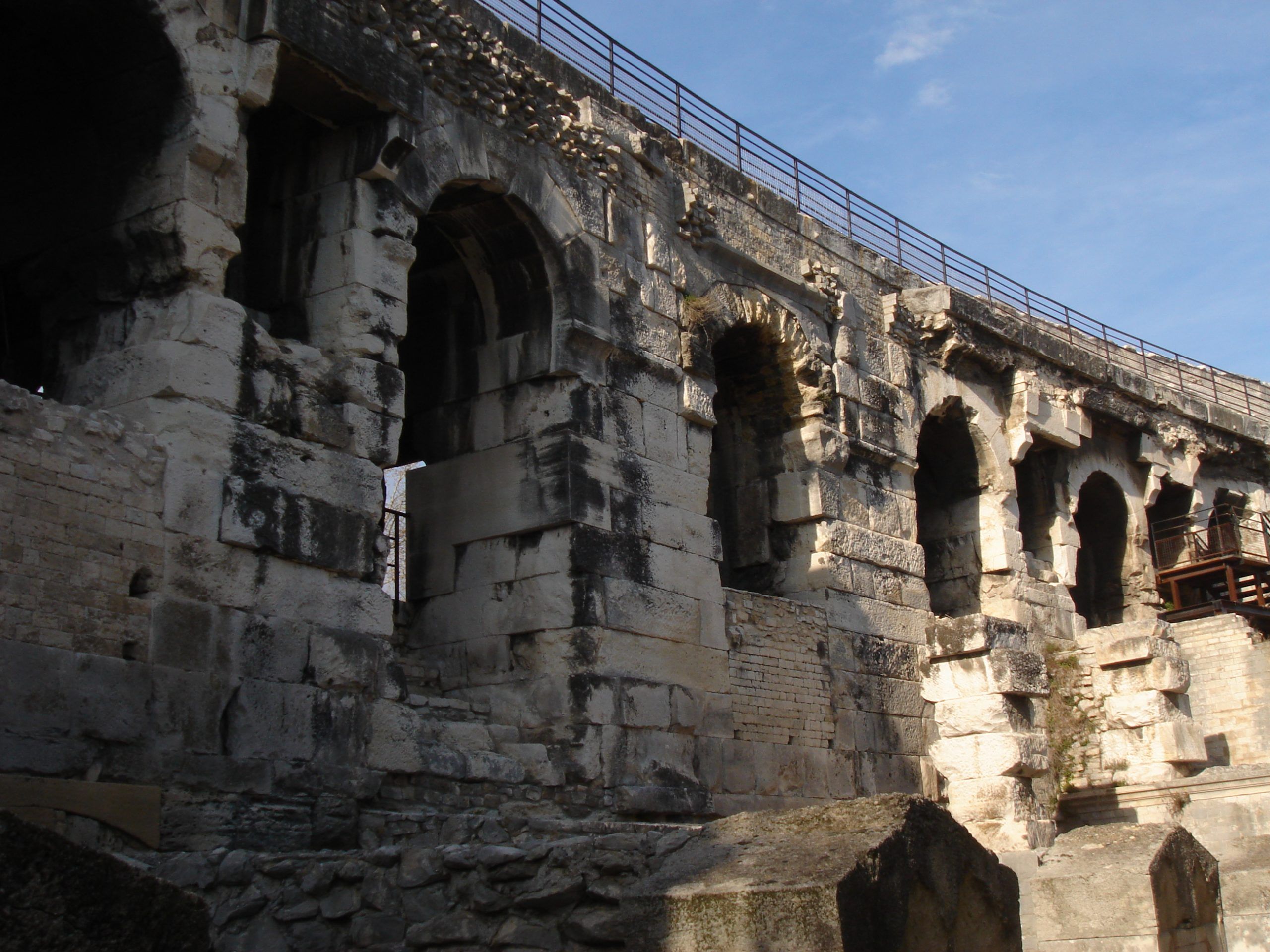 Exterior upper level of the Roman amphitheatre in Nîmes, showing weathered arches, stonework, and a modern guardrail.