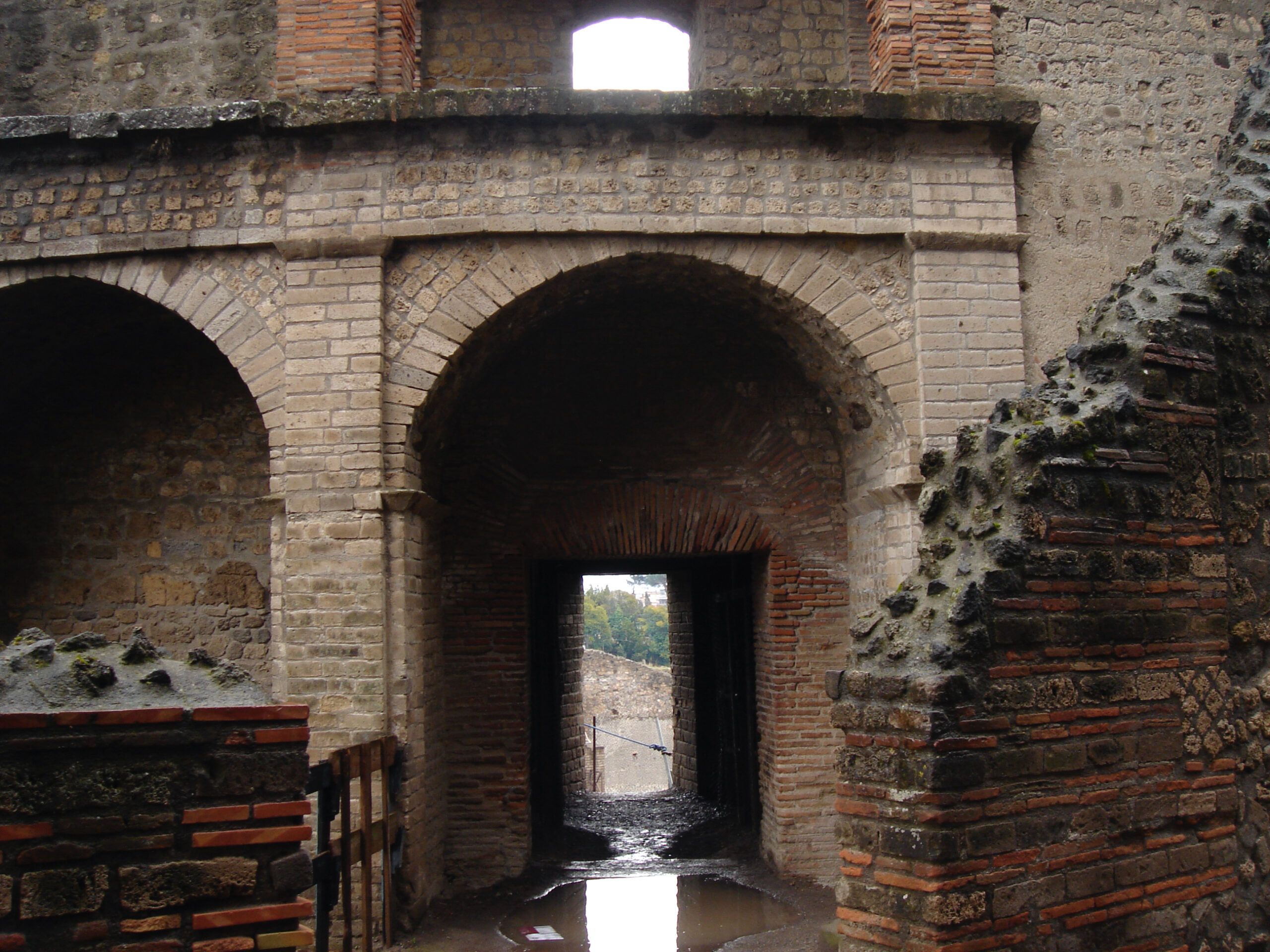 View through an arched brick passageway in Pompeii, with wet paving stones and a glimpse of ruins and greenery beyond.