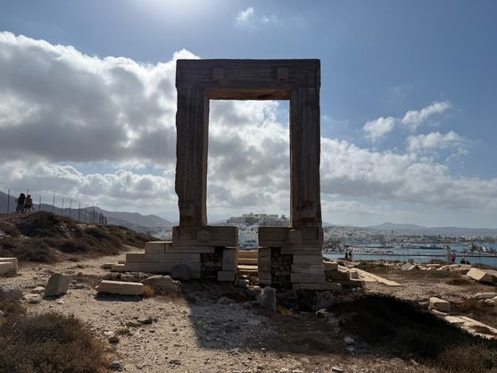 It's a marble rectangle, over two metres high, the remains of an unfinished temple. It stands on a rocky hillside on the coast; it's surrounded by marble blocks. Through it the white blocky buildings of the town of Naxos are visible, as well as  dark, sunlit clouds.