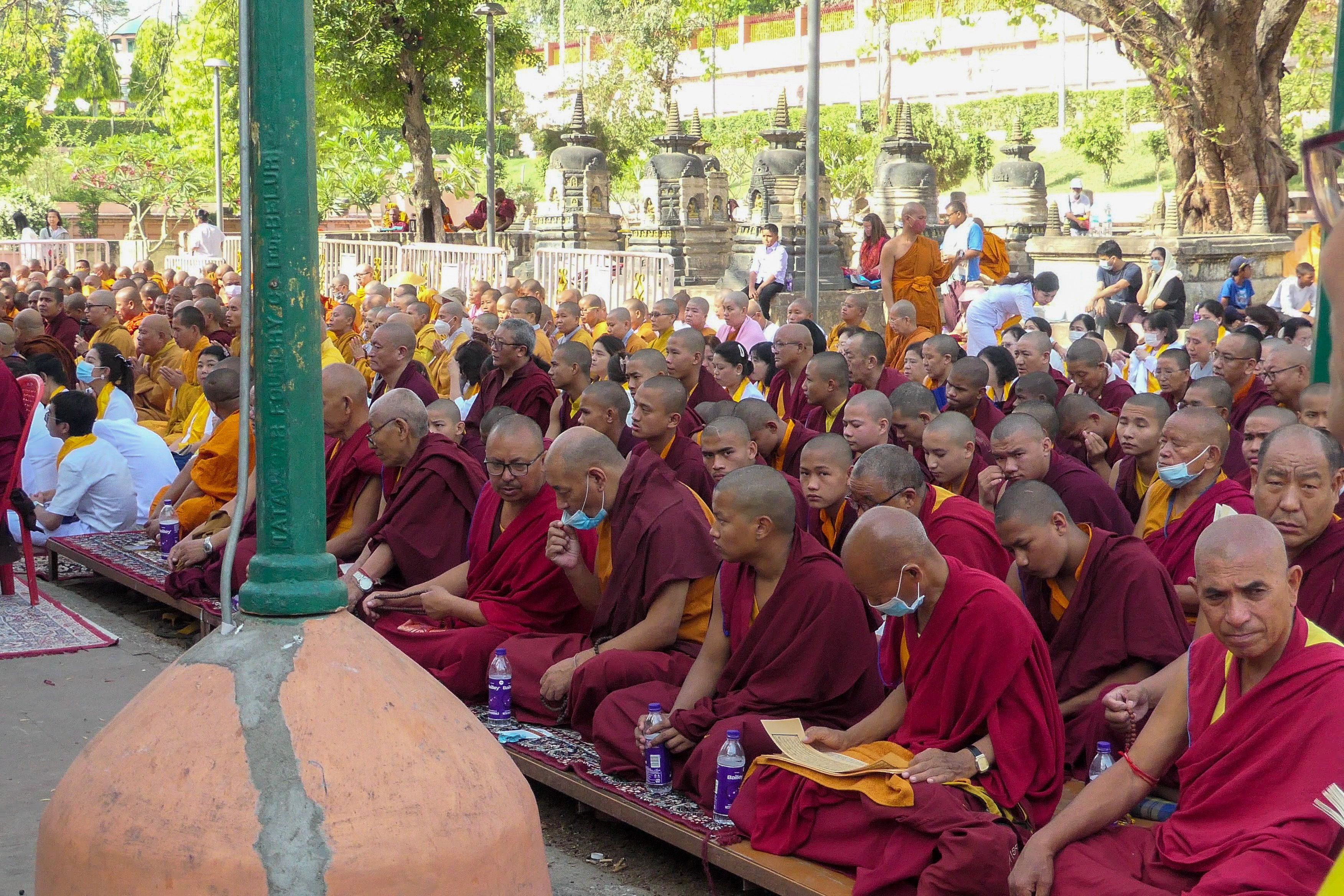 A large gathering of monks in red and orange robes sit and chant in an outdoor courtyard during a religious event.