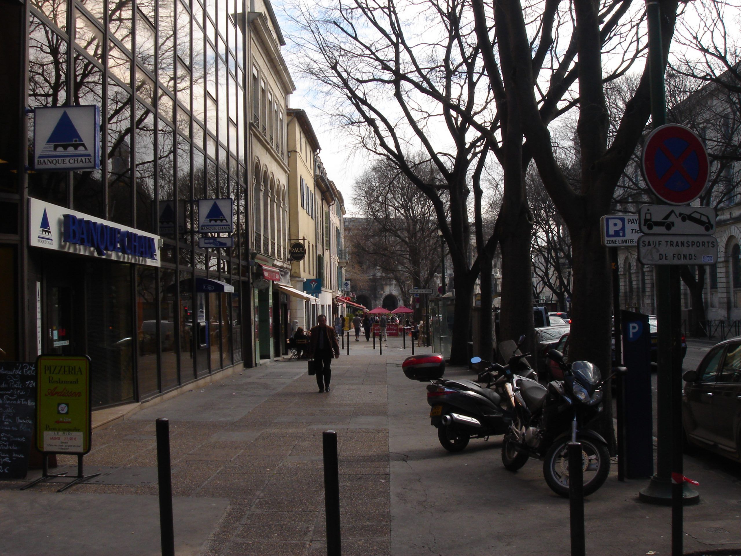 Streetscape in Nîmes with pedestrians, shopfronts, parked scooters, and a Banque Chaix branch.