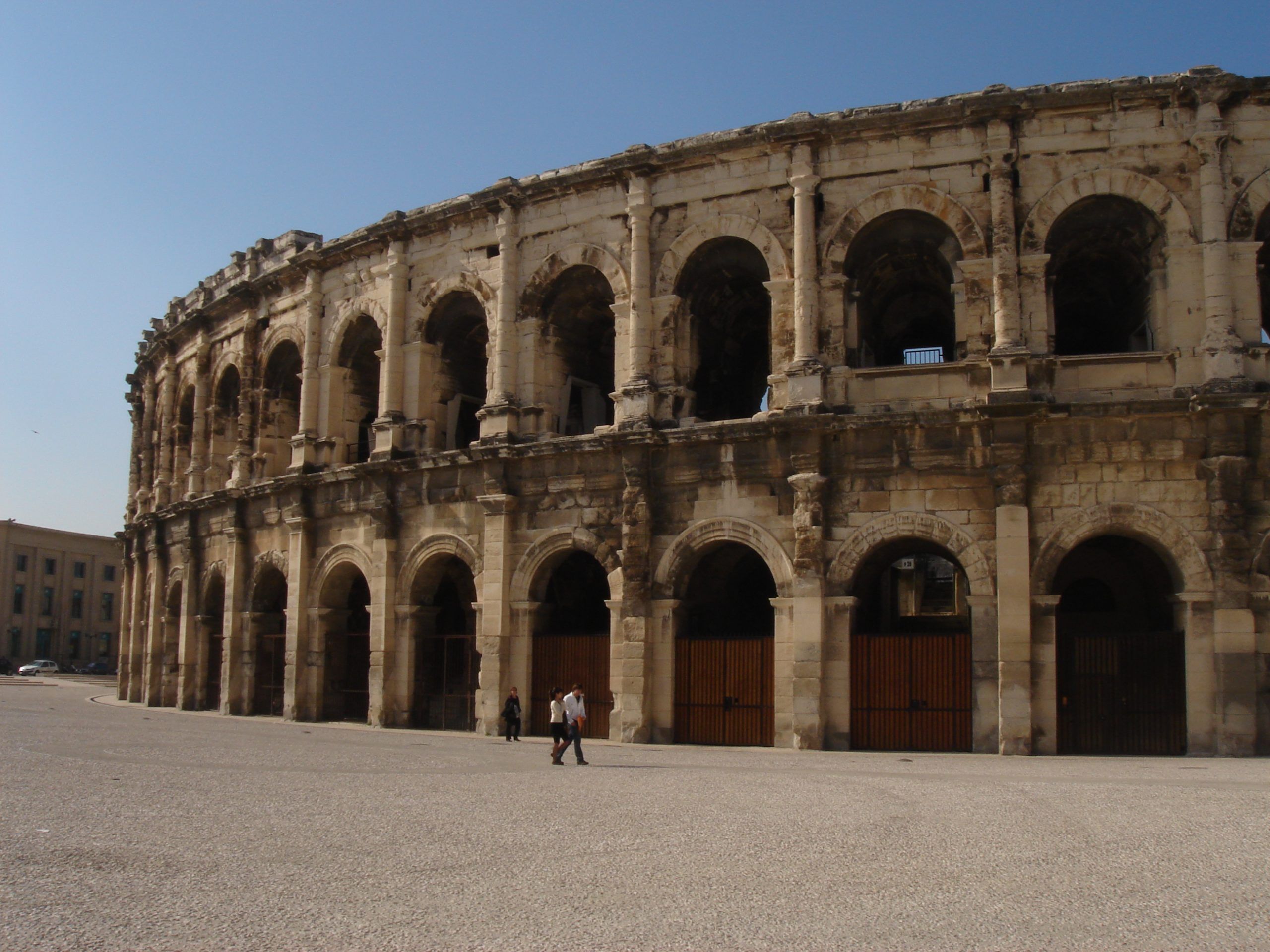 Exterior view of the Roman amphitheatre in Nîmes, with arched tiers and a mostly empty plaza.