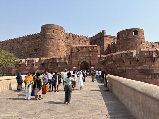 Tourists approaching the grand red sandstone gateway of Agra Fort, with its massive round bastions and crenellated walls under a clear midday sky.