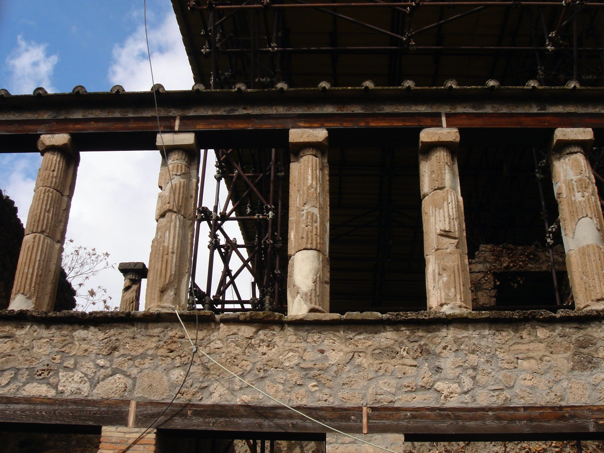 Partially restored upper floor of a Pompeian house with damaged fluted columns and extensive scaffolding behind the colonnade.