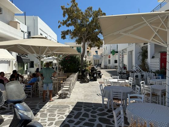A street in Paros. There are white buildings on either sides, outdoor restaurant seats under umbrellas. The sun is beating down from a blue sky.