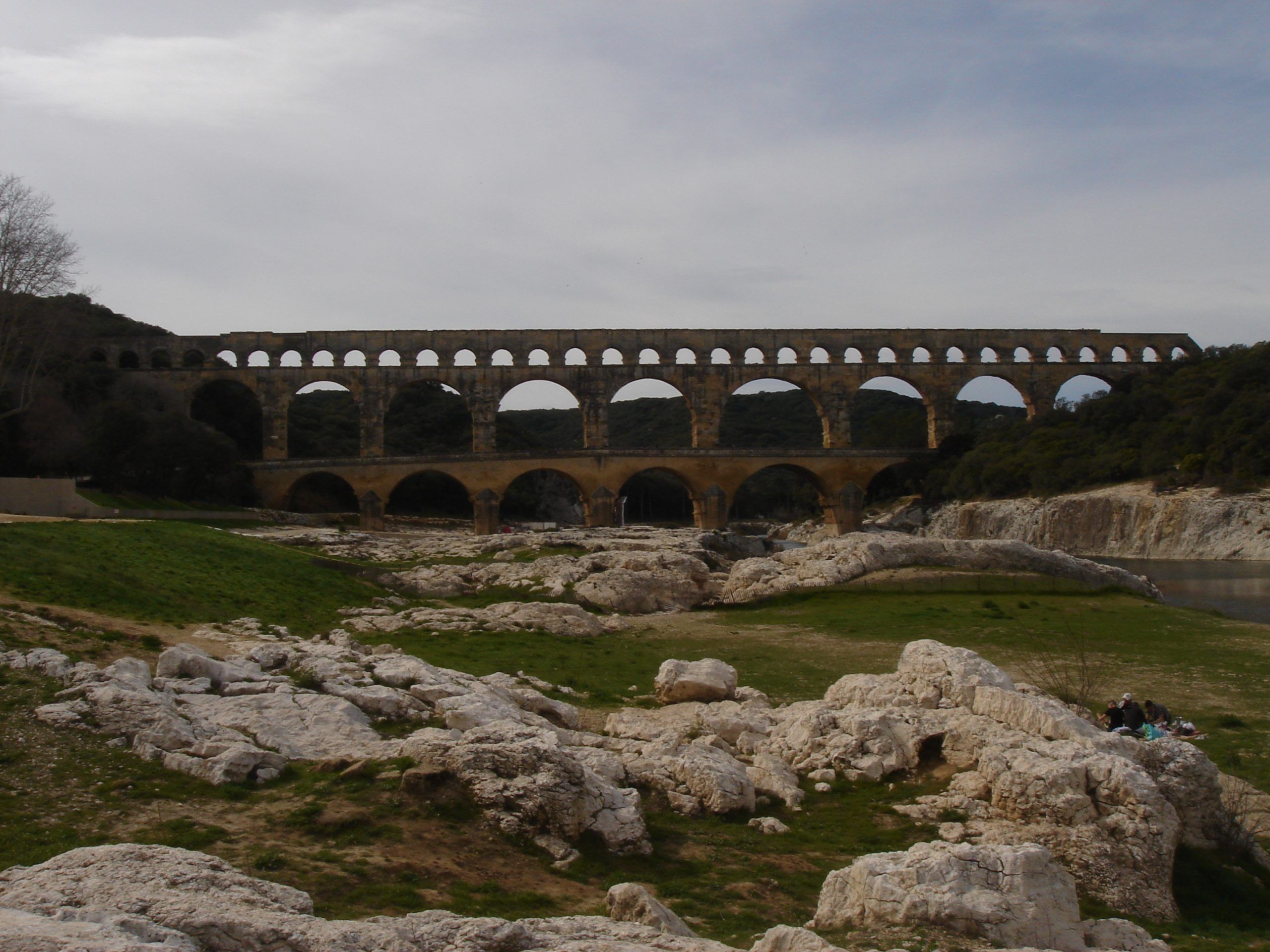 Wide view of the Pont du Gard aqueduct spanning a rocky riverbed, with green grass and limestone boulders in the foreground.