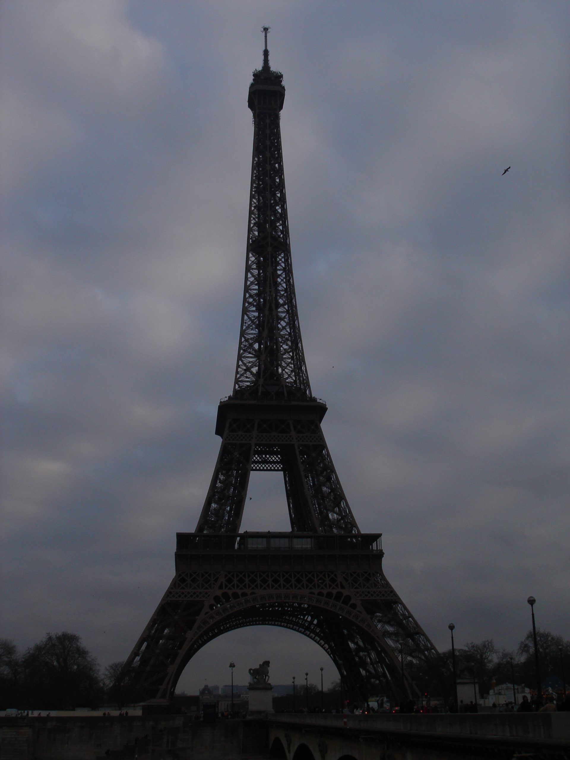 Full view of the Eiffel Tower under cloudy skies, seen from across the Seine River.