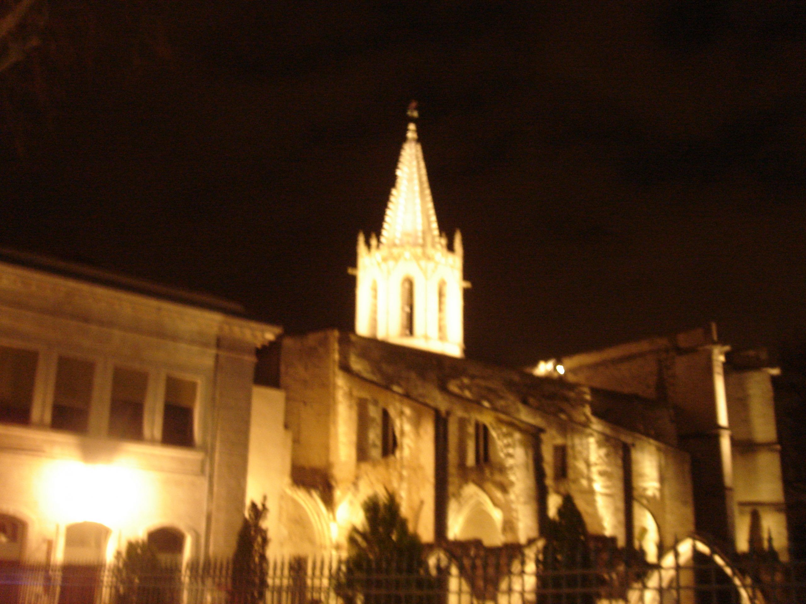 Illuminated church spire in Avignon glowing against the night sky, surrounded by darkened buildings.