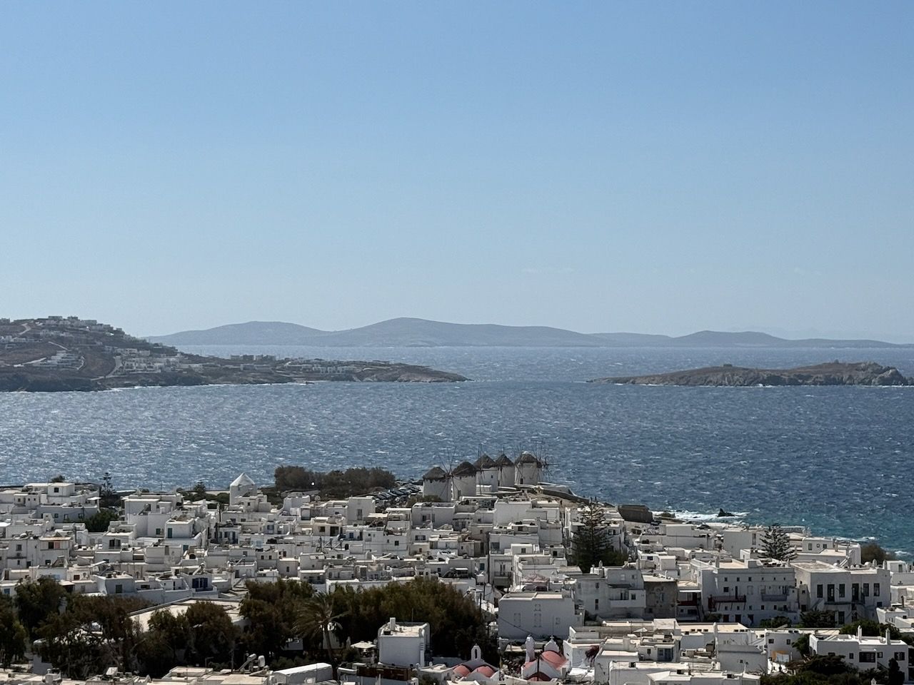 Looking out over the old town of Mykonos. In the background, the edges of the bay and an island in the distance. At the edge of the water, a row of cylindrical windmills. Then a jumble of low white flat-roofed buildings.