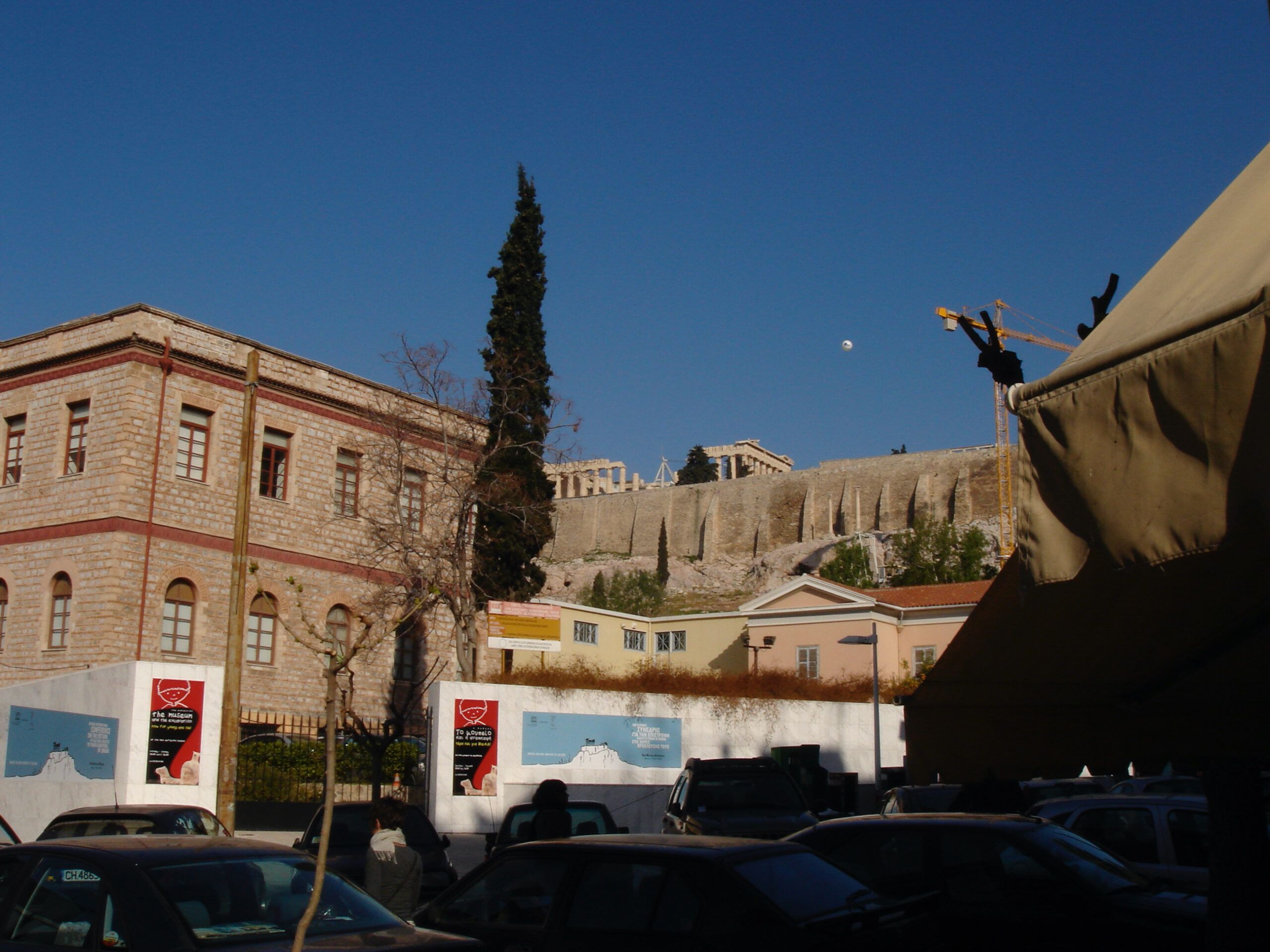 View of the Acropolis in Athens from a nearby street, with the Parthenon visible above the city wall and modern buildings in the foreground, including construction cranes and a canvas awning.