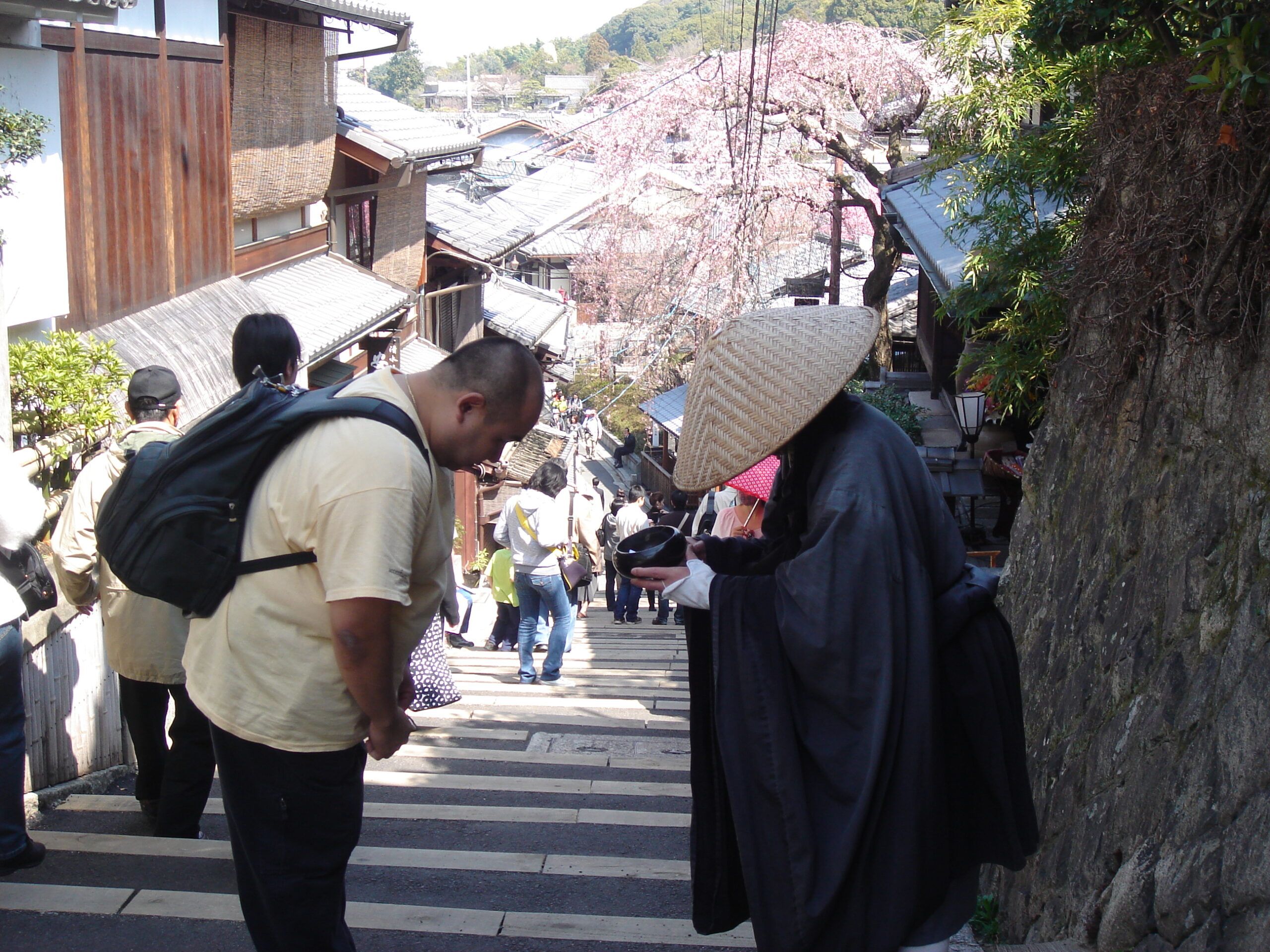 Calvin bows respectfully to a monk in black robes and a straw hat, who is standing on a stone staircase lined with traditional wooden buildings and cherry blossoms.