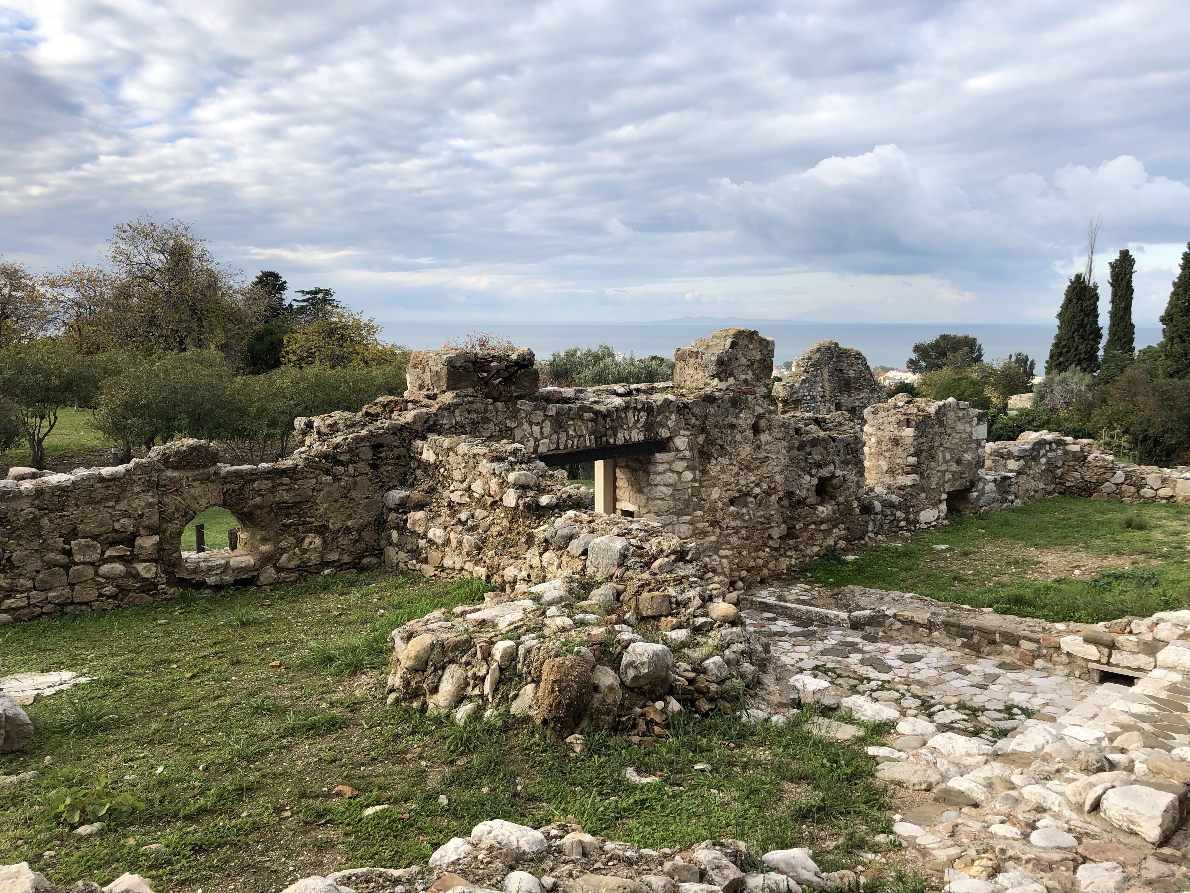 Low walls, one with an arch and the remains of flooring and internal  walls. Most of the ground is grass and there are trees outside.